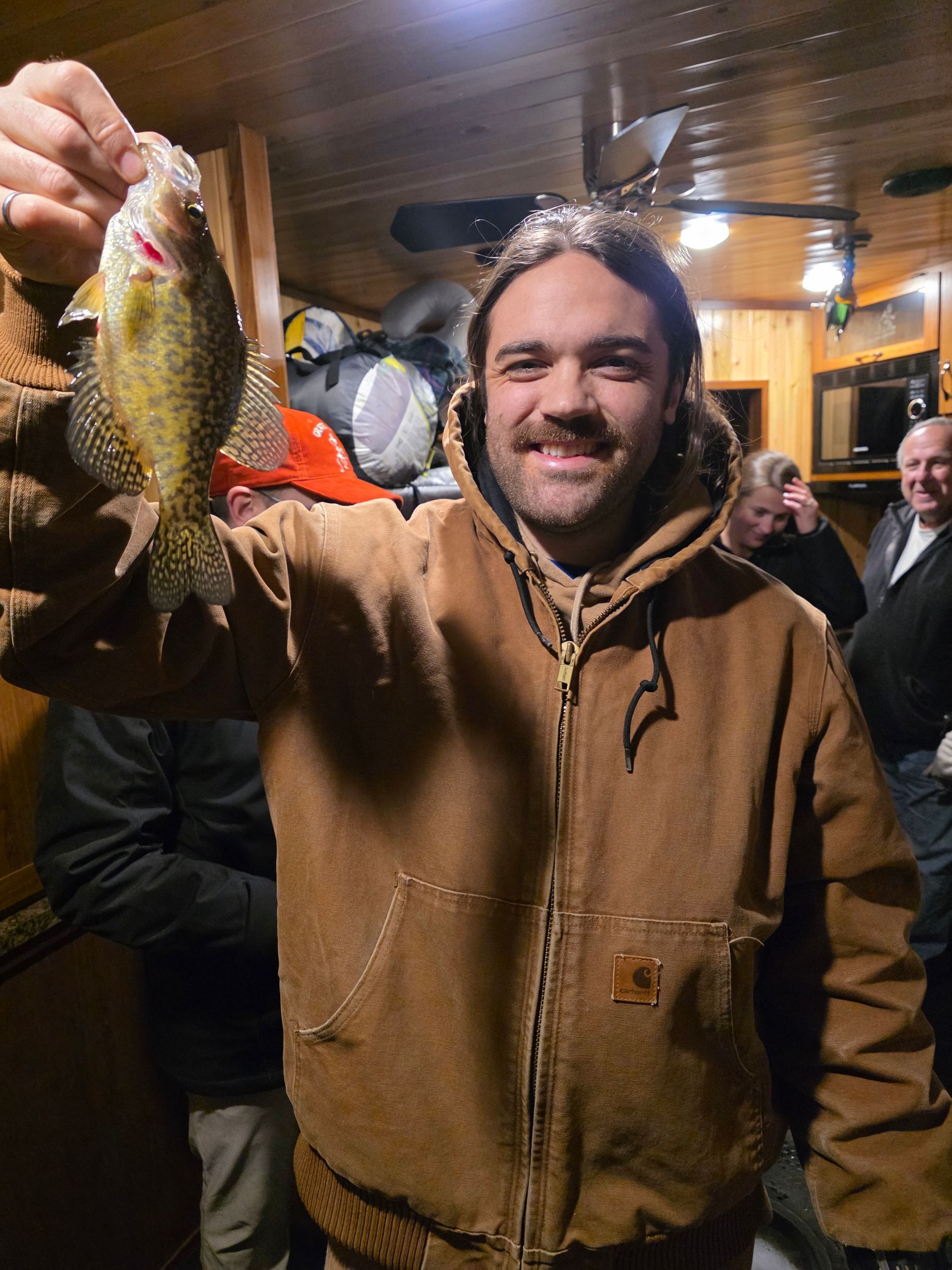 Man in brown jacket smiling and holding up a fish indoors at a lively gathering