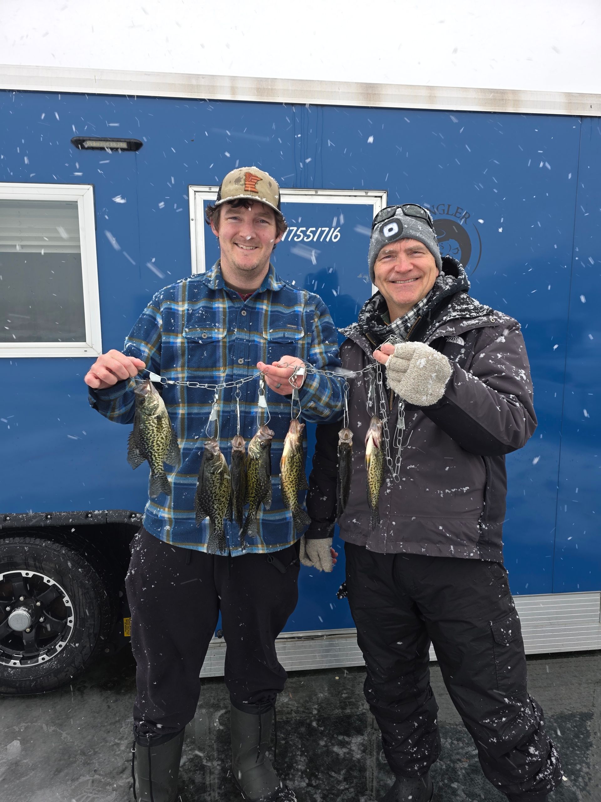 Two people in winter gear smiling and holding fish in the snow beside a blue truck