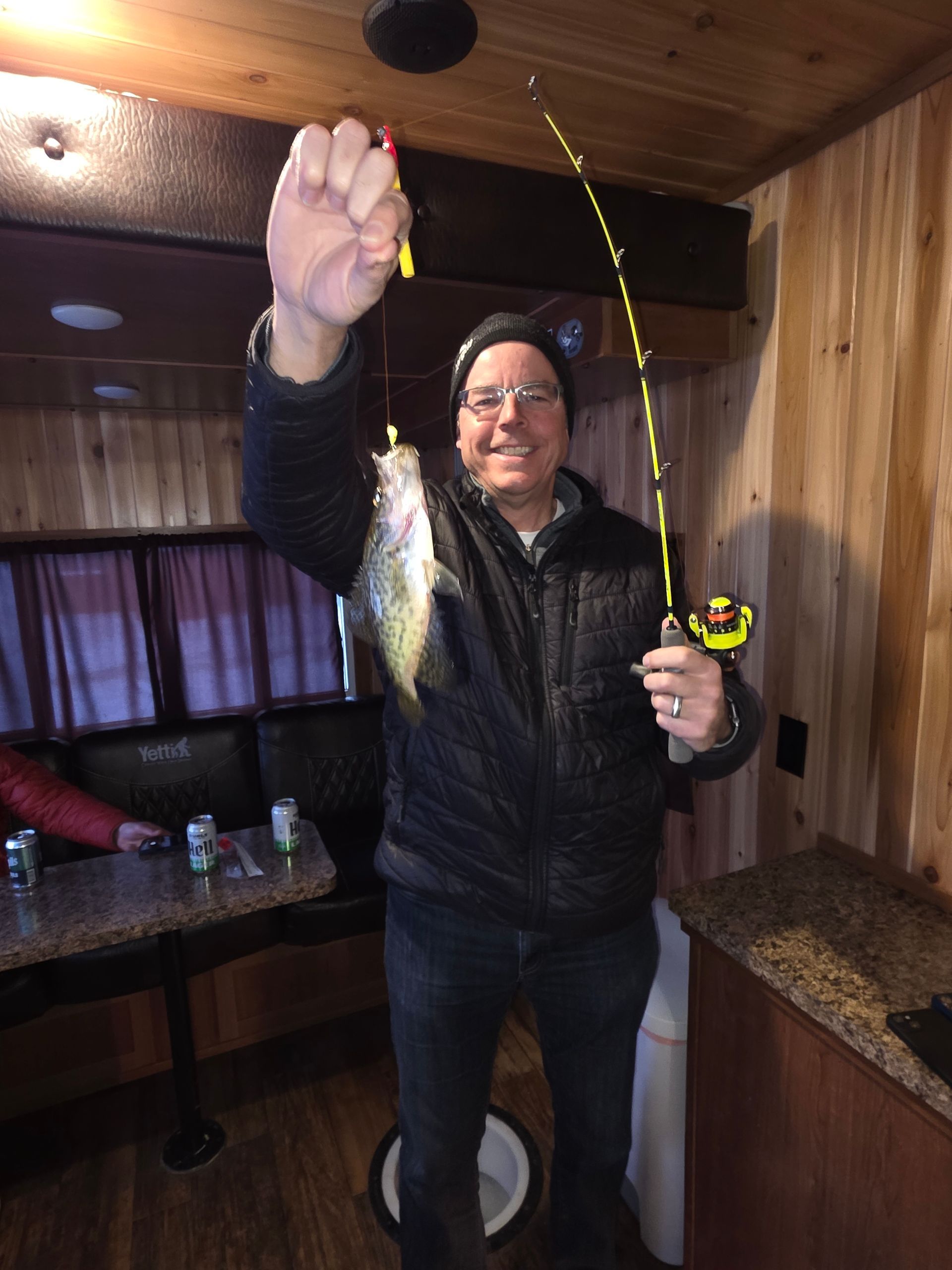 Person indoors holding a fishing rod and a fish, smiling in a wood-paneled room.