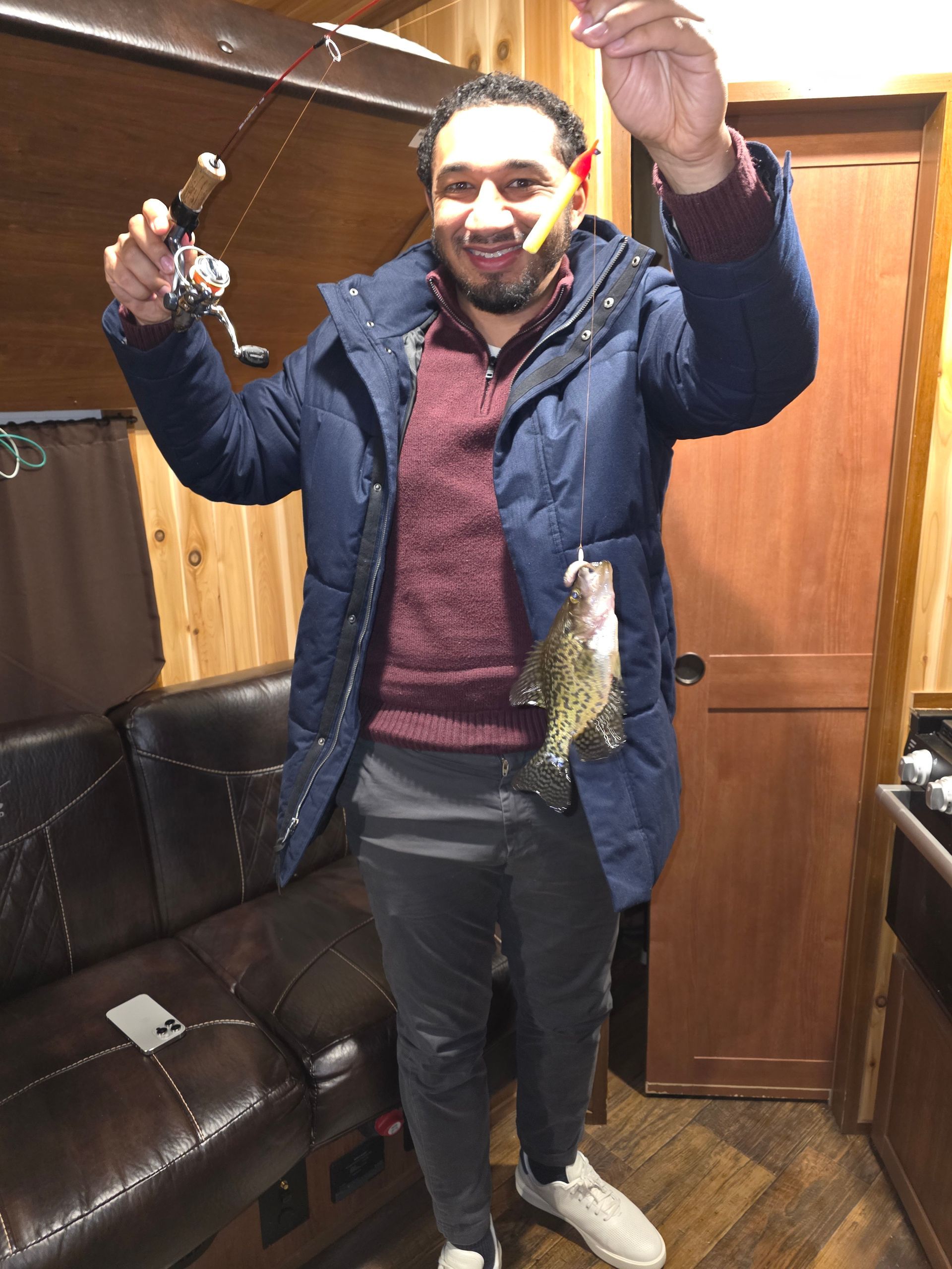 Man indoors holding two fish, smiling in a wood-paneled room with a leather couch.
