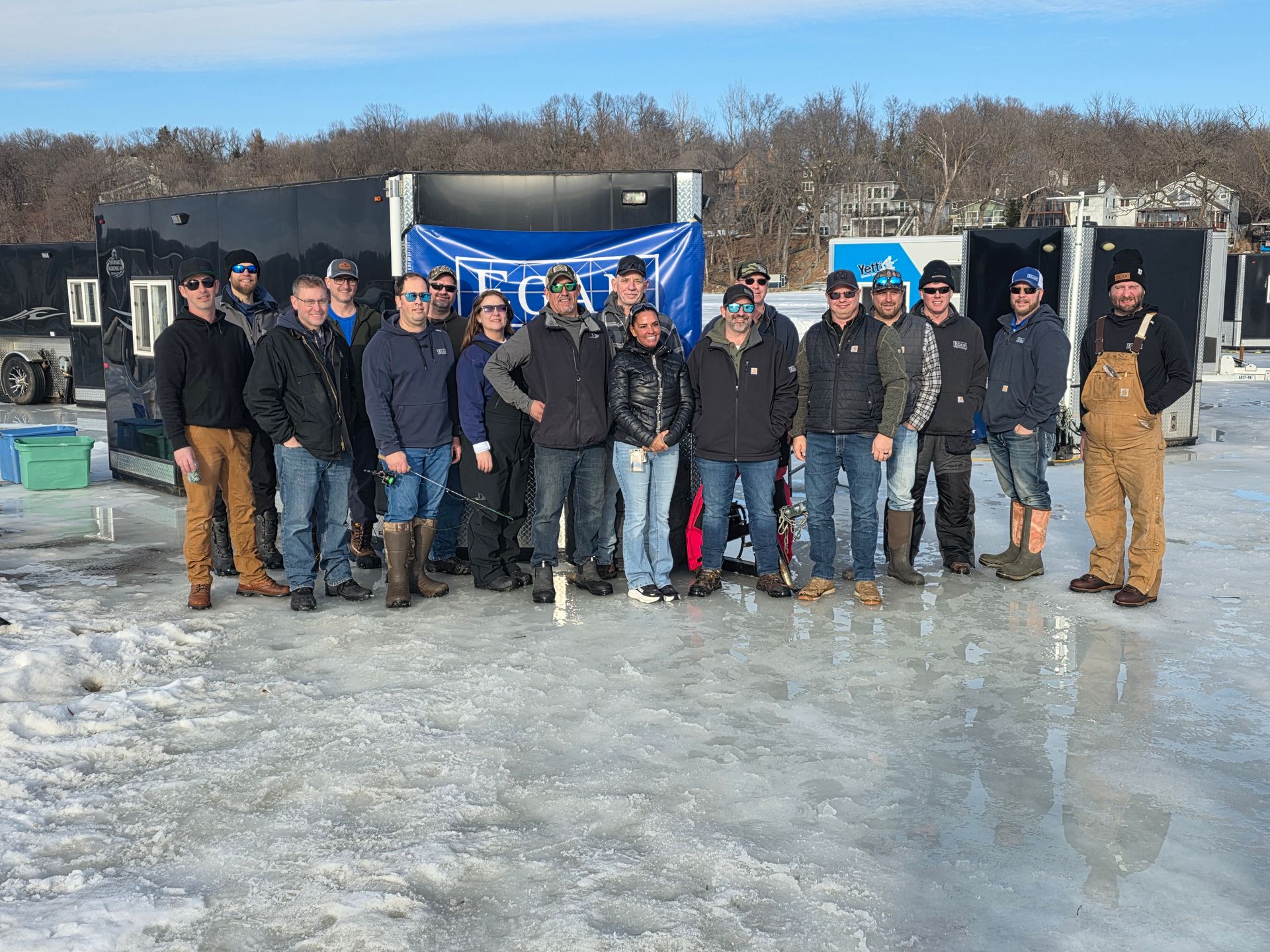 Group of people posing on an icy field in winter, standing before a blue banner and equipment trailer.