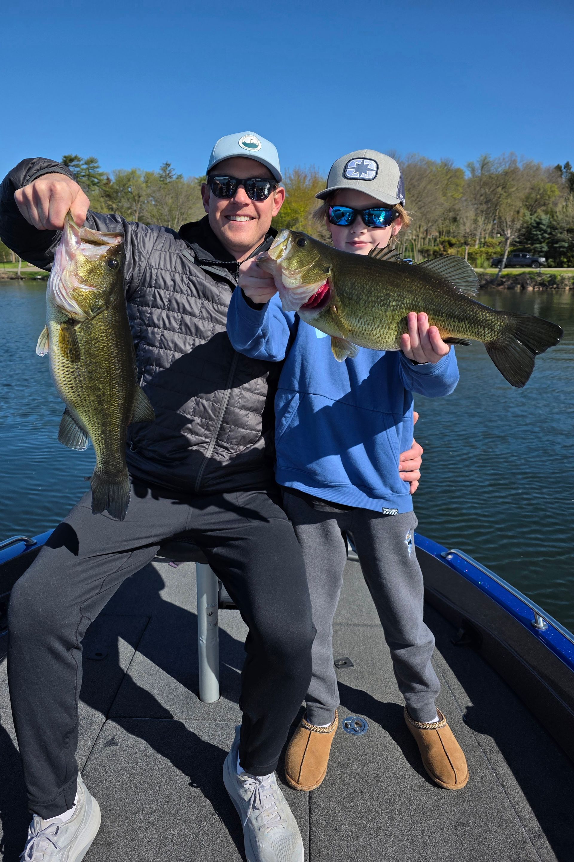Two people on a boat hold up large fish on a sunny lake, smiling in sunglasses and hats.