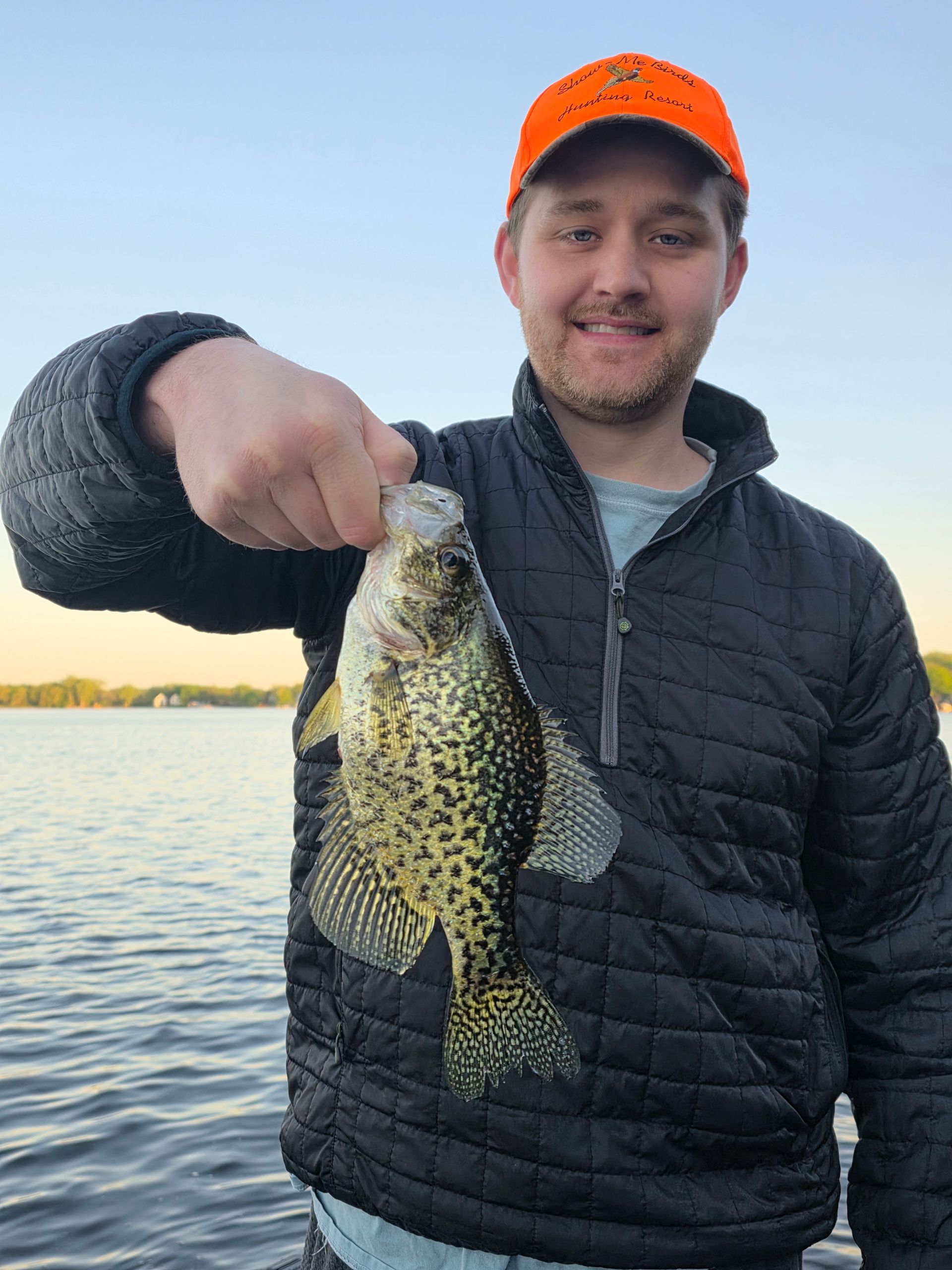 Man holding a speckled fish by the water at sunset, wearing an orange cap and black jacket.