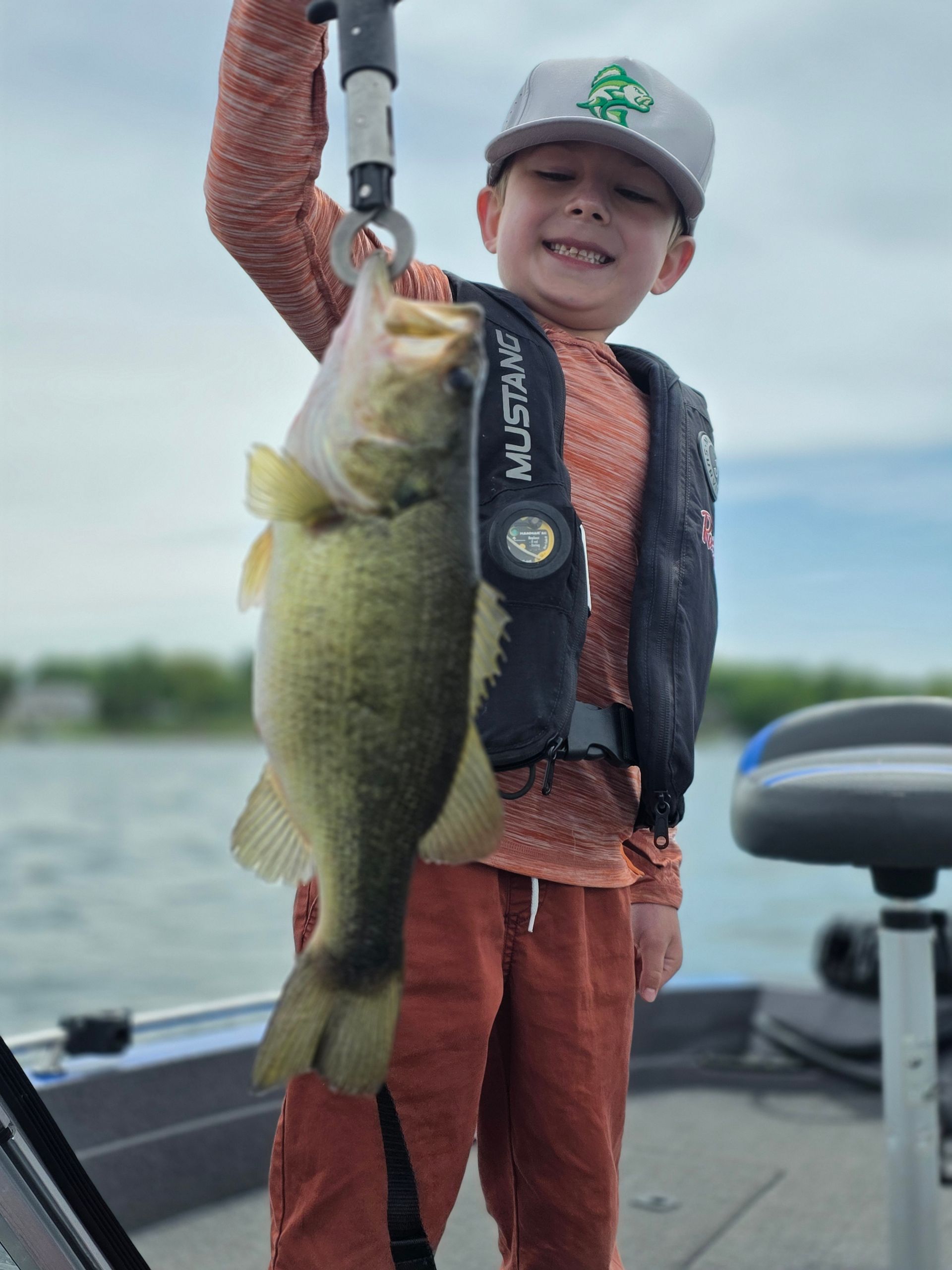 Child on a boat holding up a large fish, wearing a cap and life vest on the water.