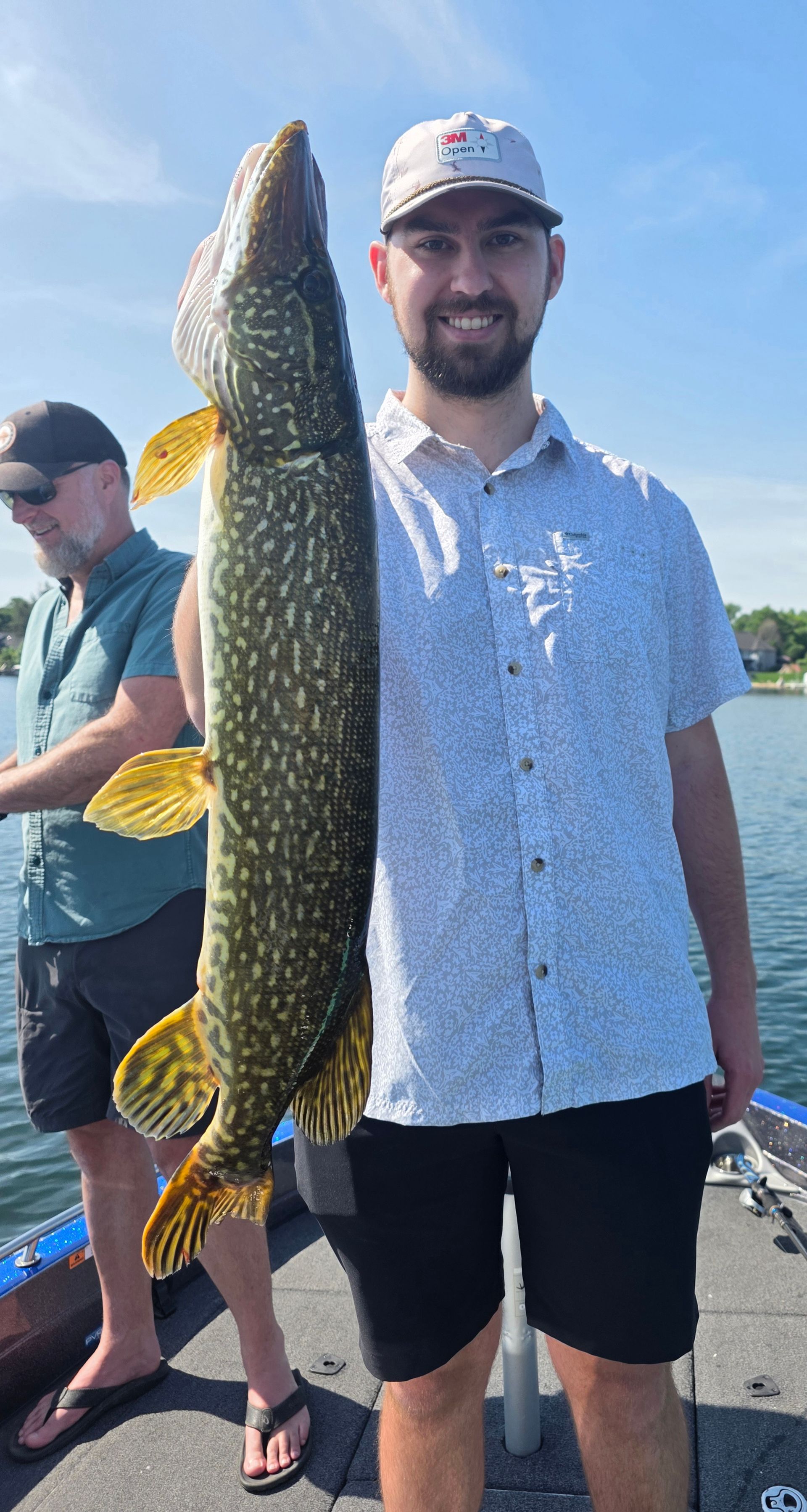 Man holding a large speckled fish on a boat, with another person and water in the background.