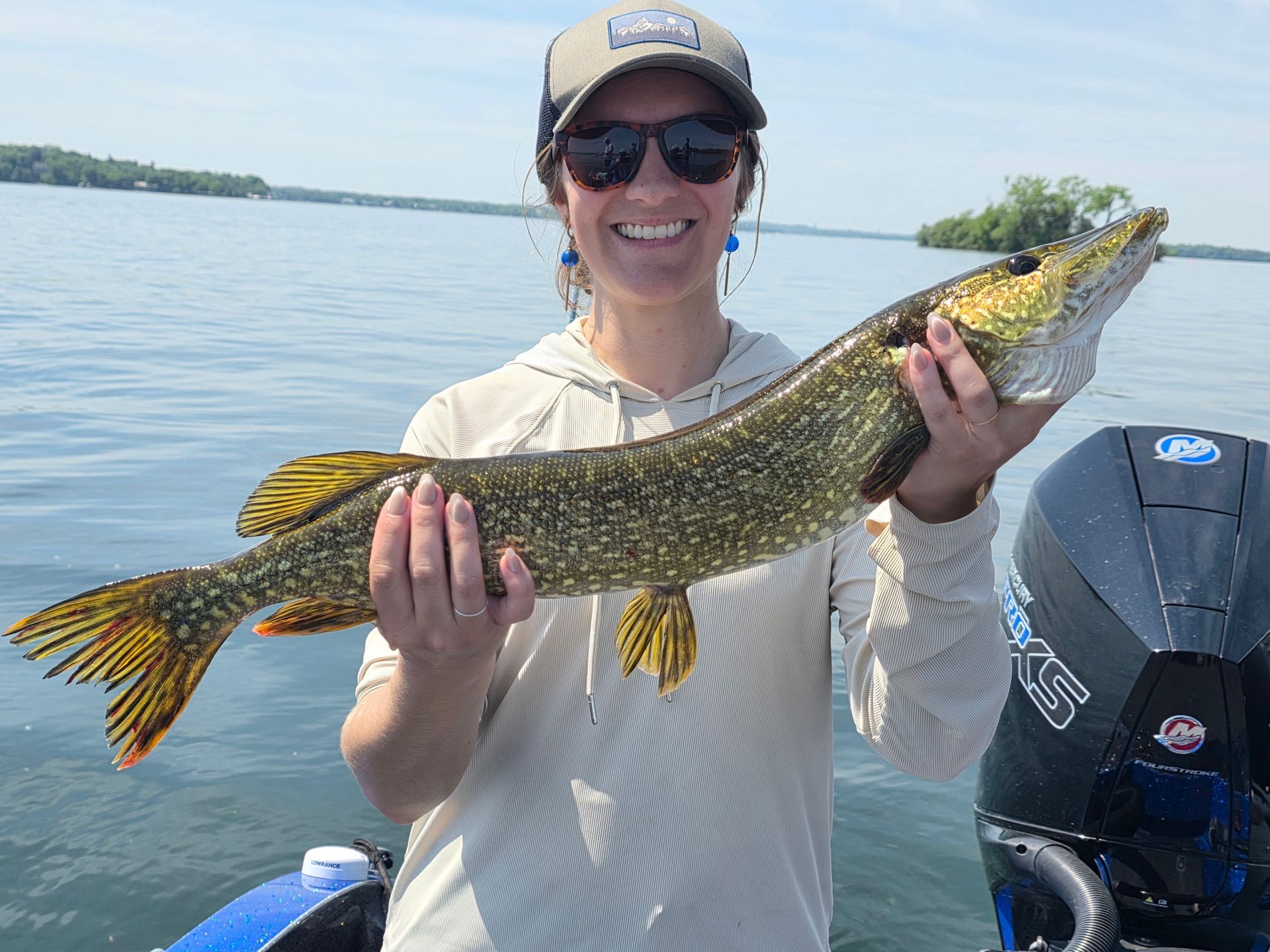 Person holding a large fish beside a boat on calm water, smiling in sunglasses and a cap.