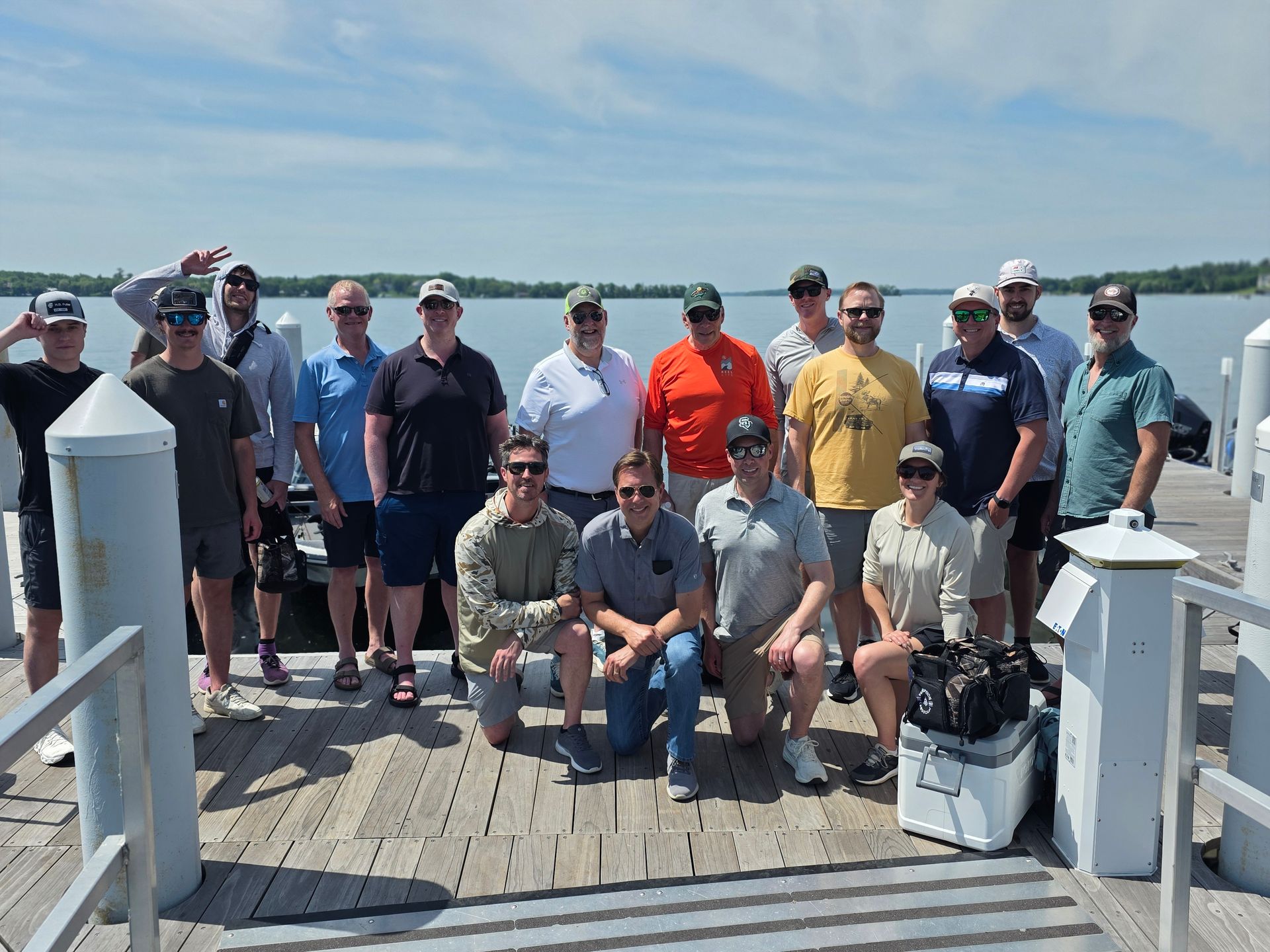 Group of people posing on a dock by the water, with blue sky and boats in the distance.