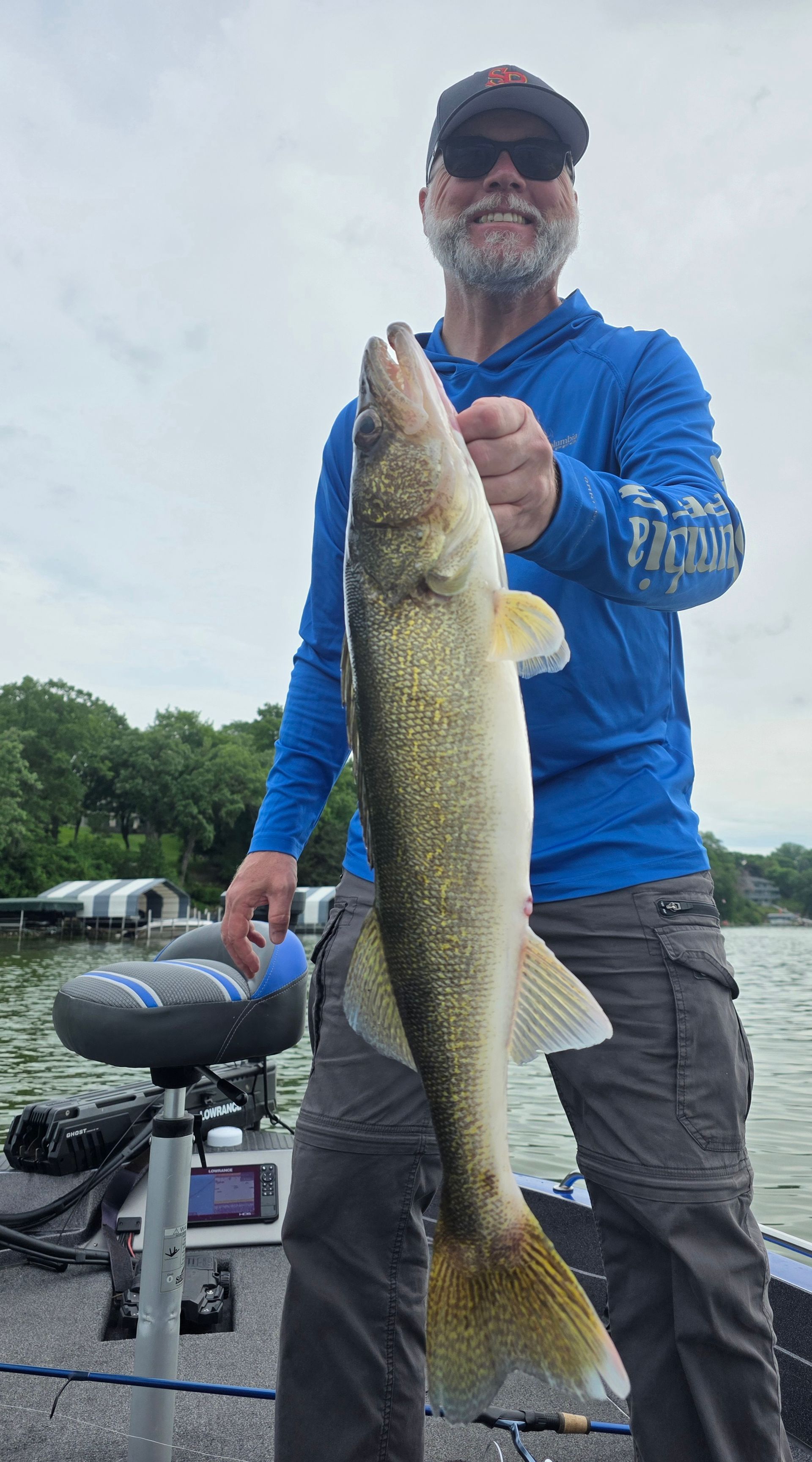 Angler in blue shirt holding a large fish on a boat with another boat and cloudy sky behind him
