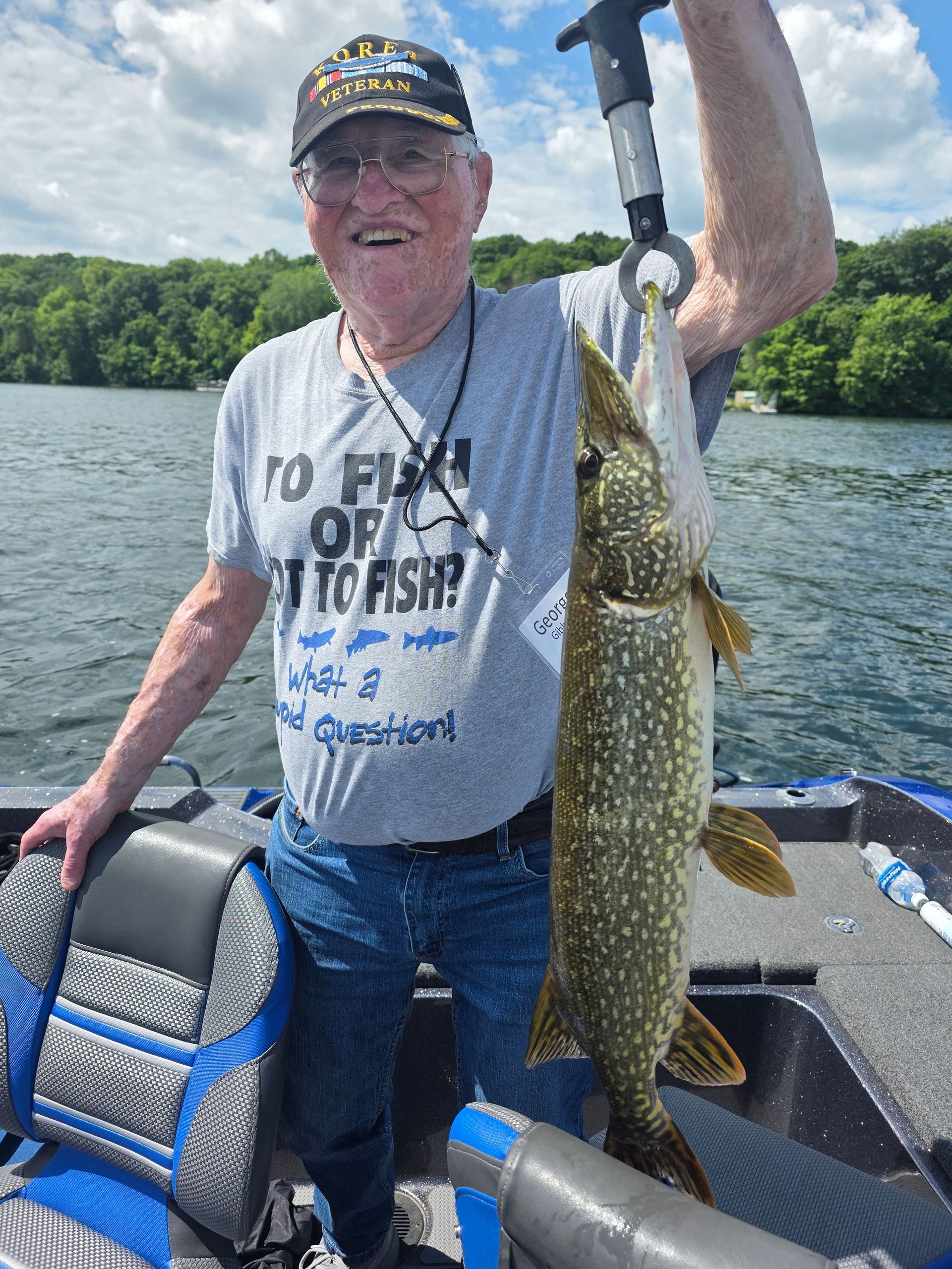 Man holding a large fish on a boat on a lake, smiling in a gray T-shirt and cap.