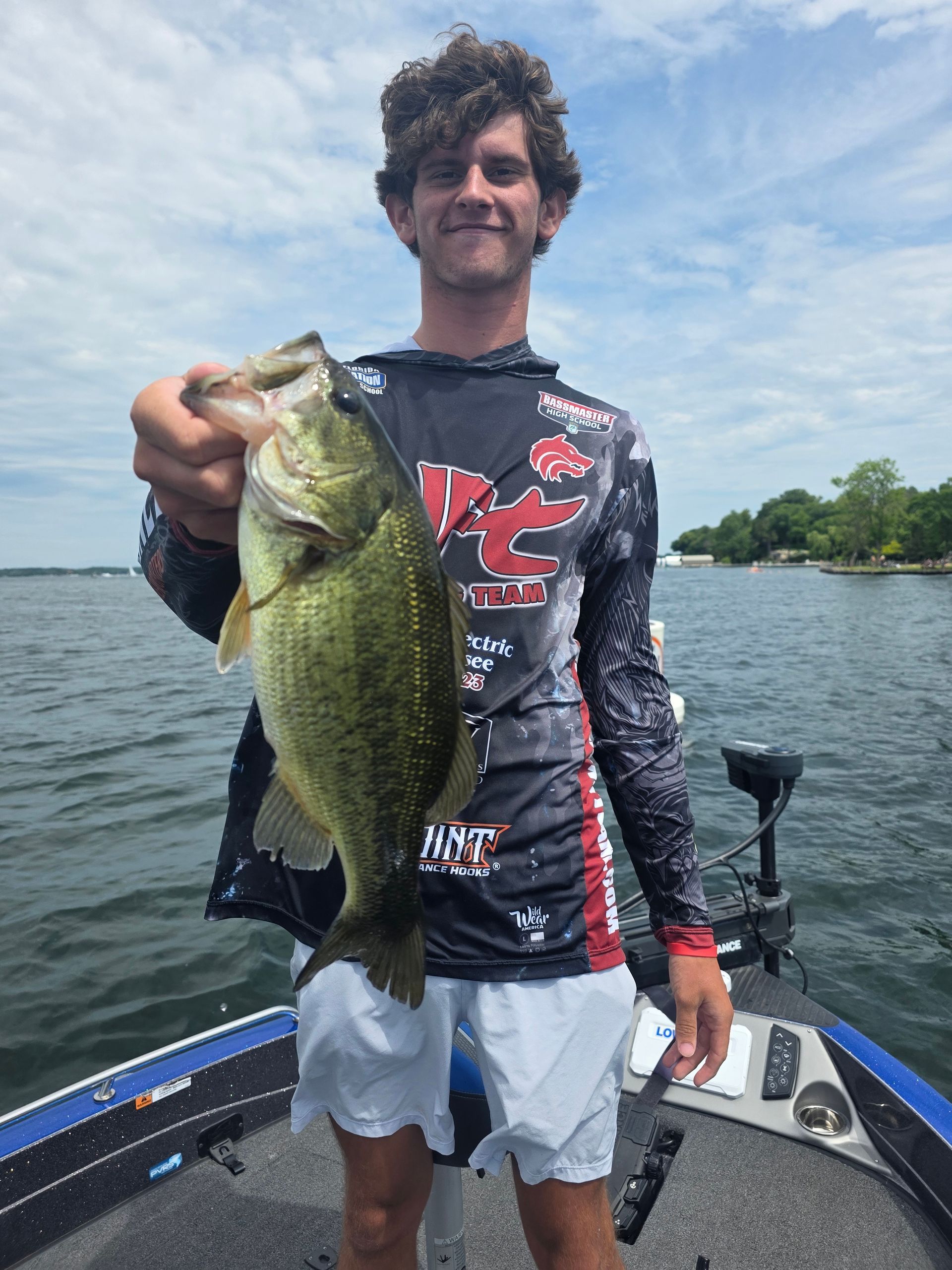 Man holding a large bass on a boat, wearing a fishing jersey on a calm lake.