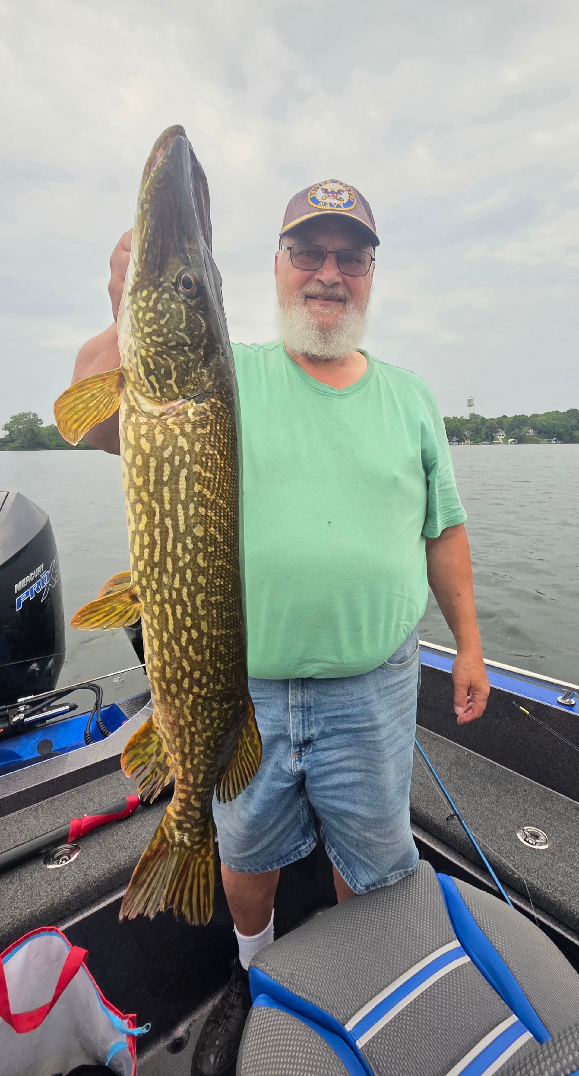 Man holding a large fish upright on a boat, wearing a green shirt and cap on calm water