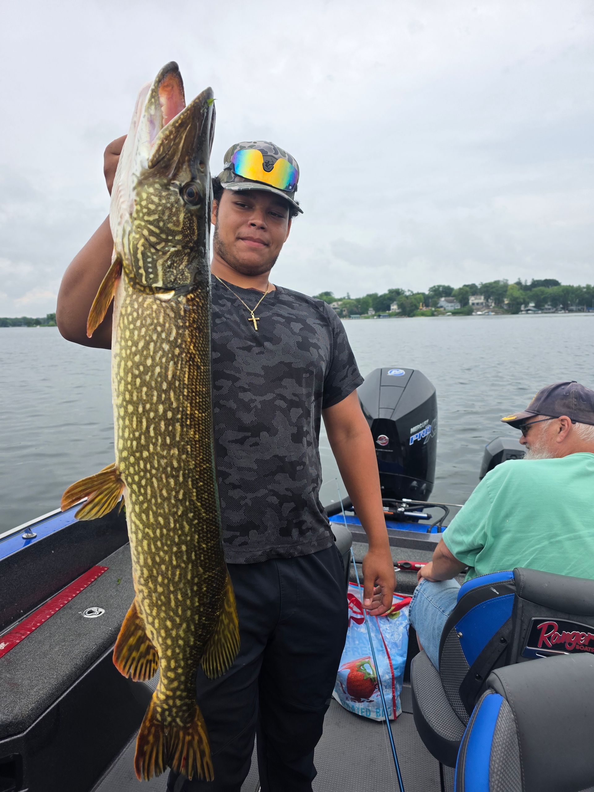 Angler on a boat holds a large pike by a lake, with another person seated nearby.