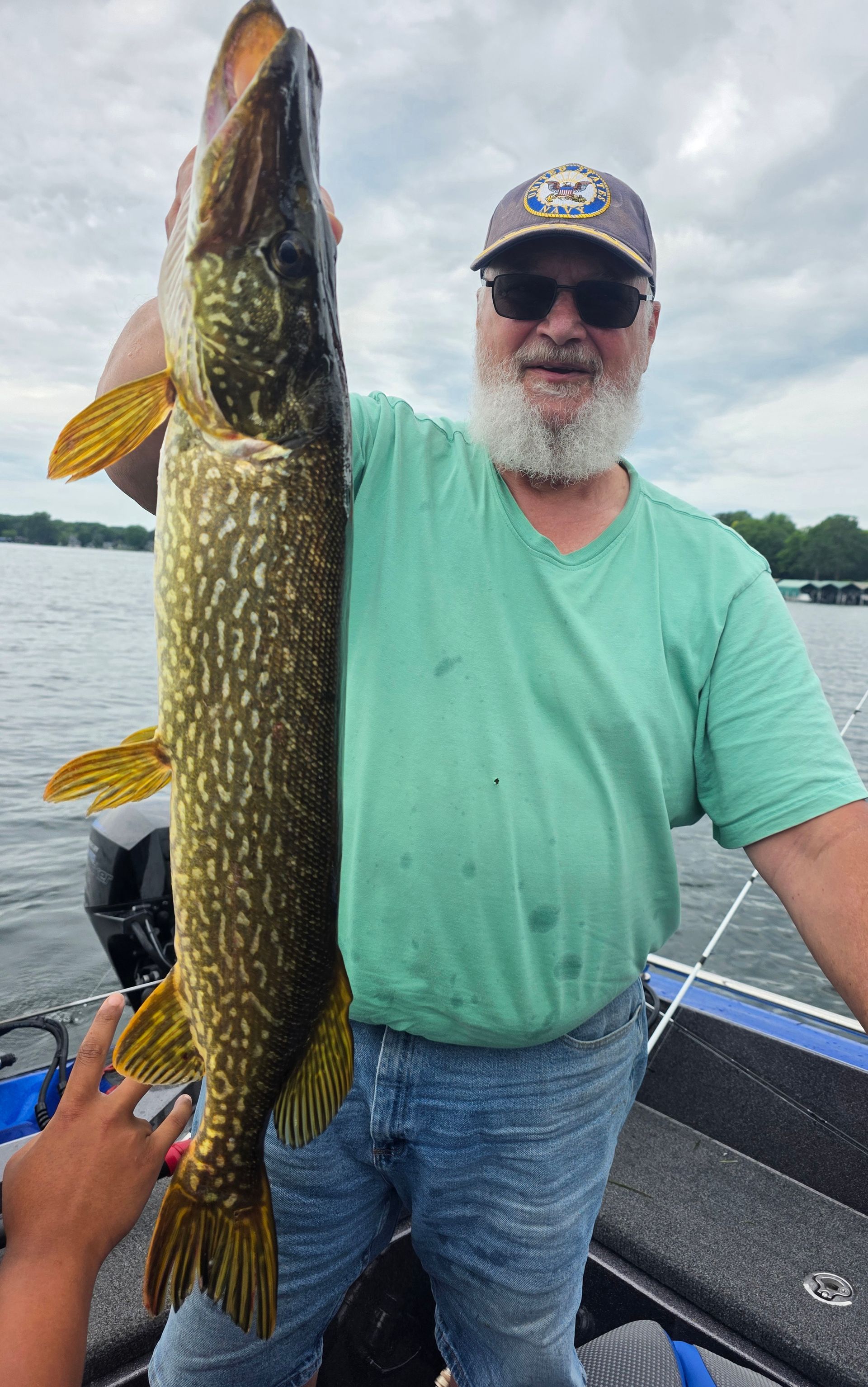 Man holding a large pike on a boat, wearing a green shirt and cap on calm water.
