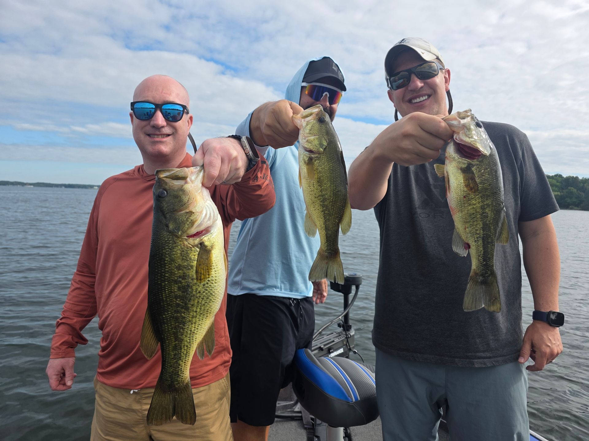 Three anglers on a boat hold up largemouth bass on a cloudy lake.
