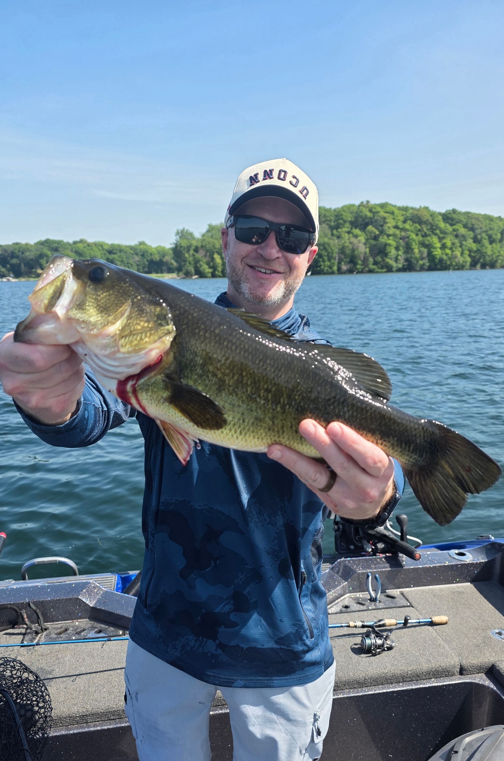 Smiling angler holding a large bass on a boat on a lake, with trees and blue sky behind him
