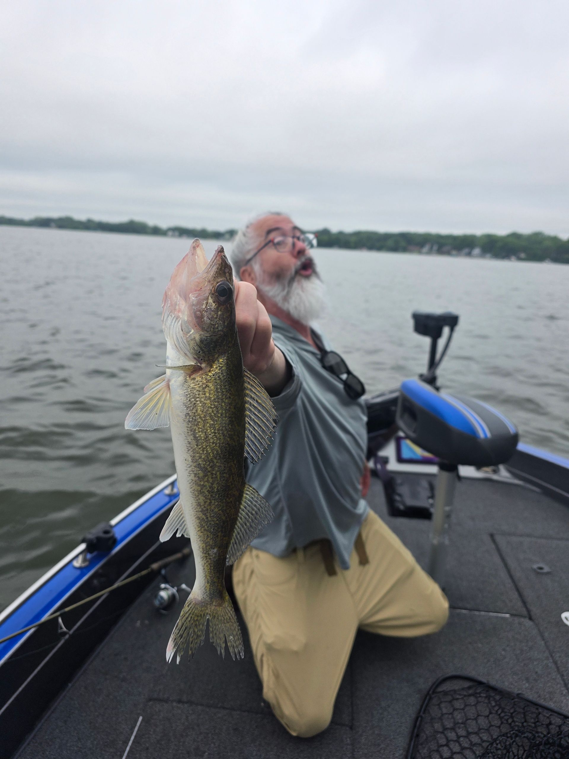 Angler kneels on a boat, holding up a large fish on a lake under an overcast sky.