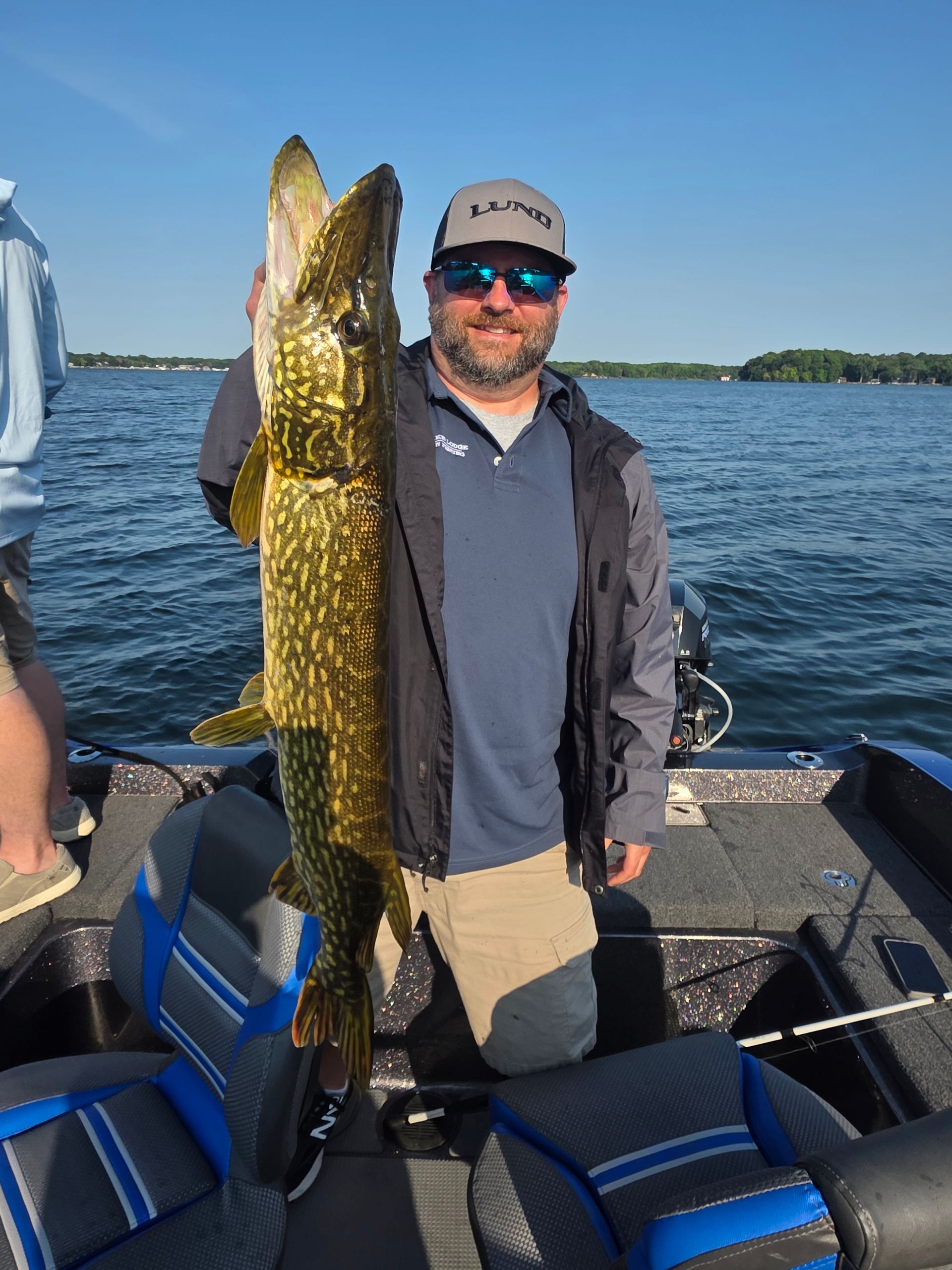 Man holding a large fish on a boat on a sunny lake, wearing a cap and sunglasses.