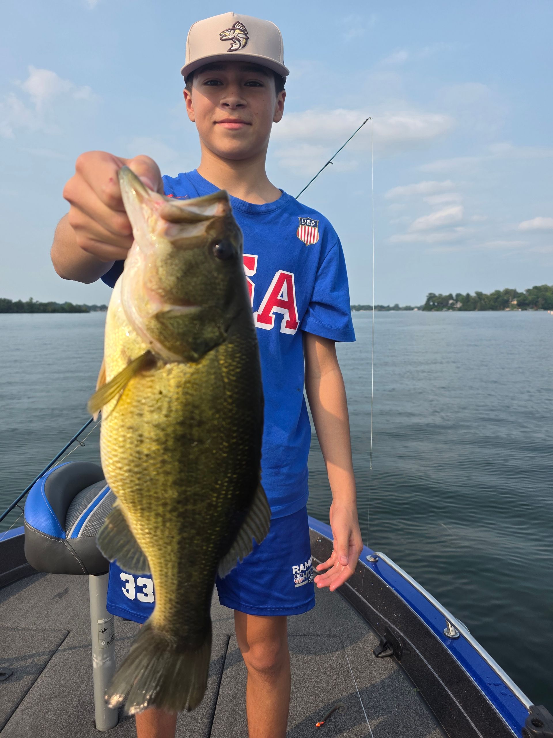 Youth holding a large bass on a boat dock over calm water, wearing a blue shirt and cap