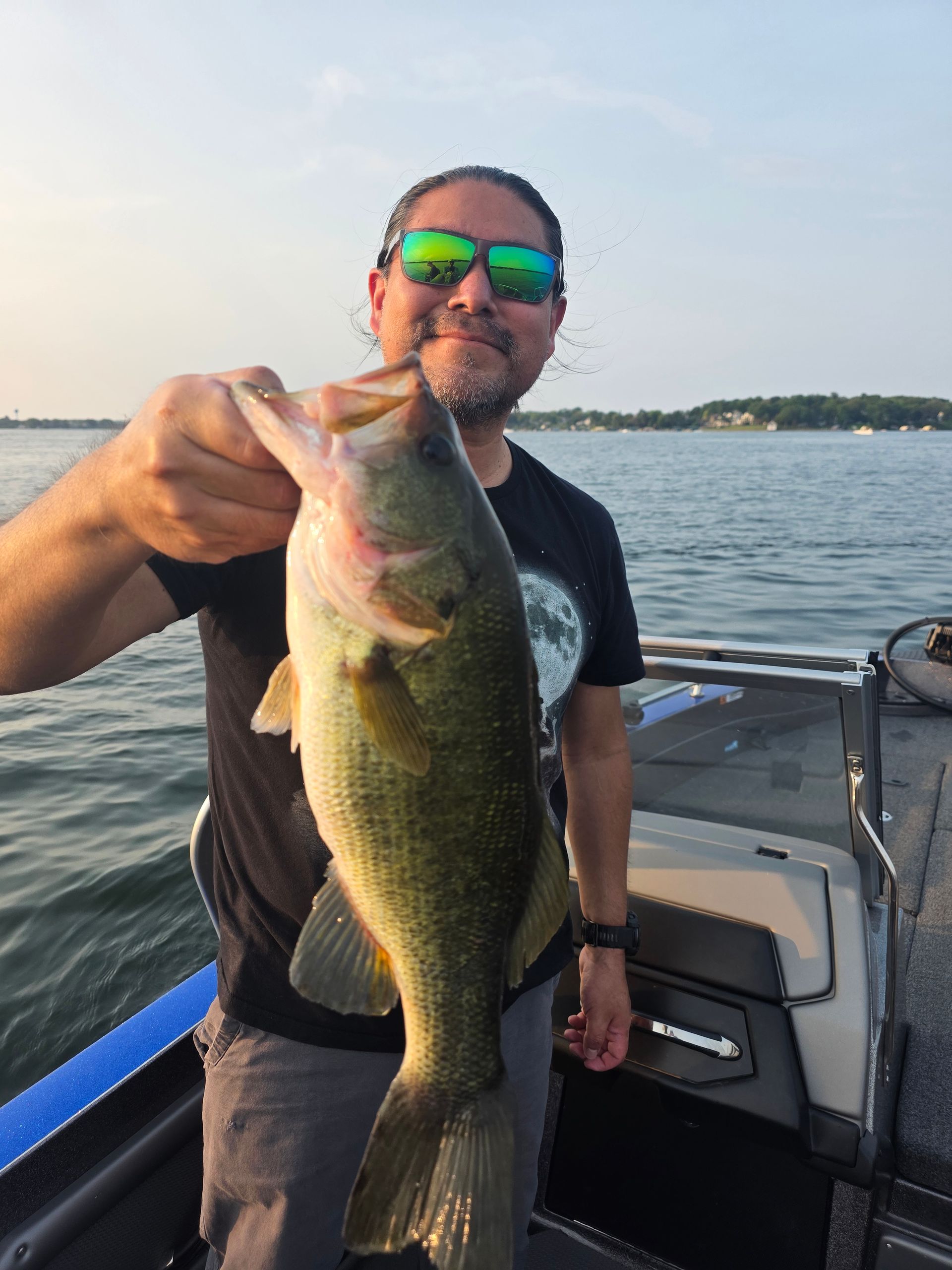 Man holding a large bass on a boat at sunset, wearing sunglasses near calm water.