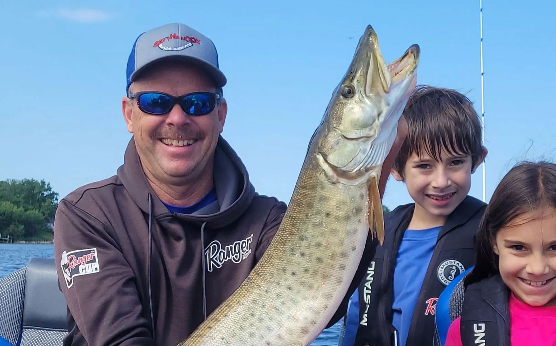 Man holding a large fish with two smiling children on a boat under a clear blue sky
