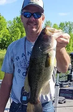 Man holding a large fish outdoors on a sunny day near a boat and trees