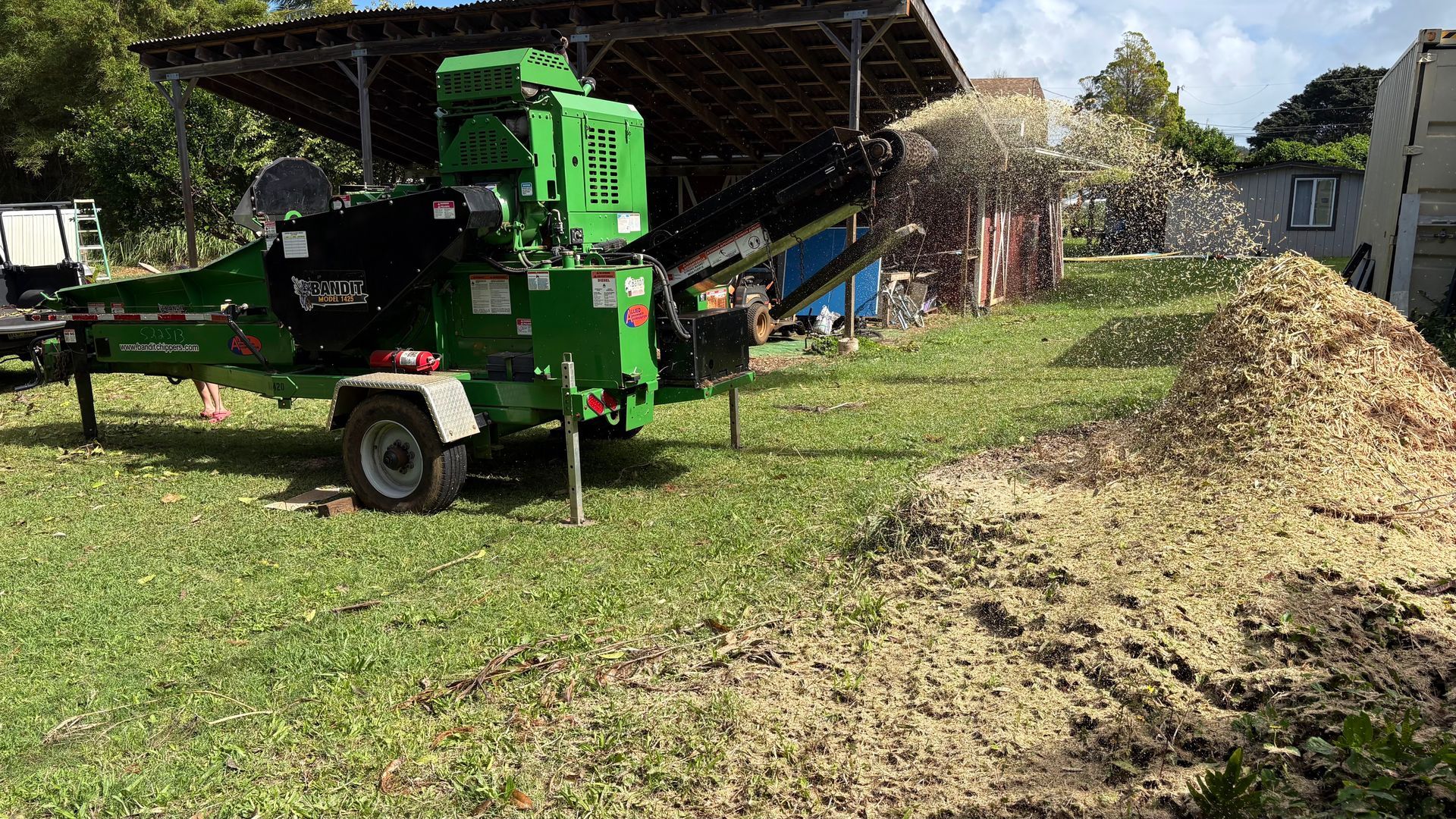 A bright green wood chipper machine in a grassy yard, spraying wood chips into a large pile.