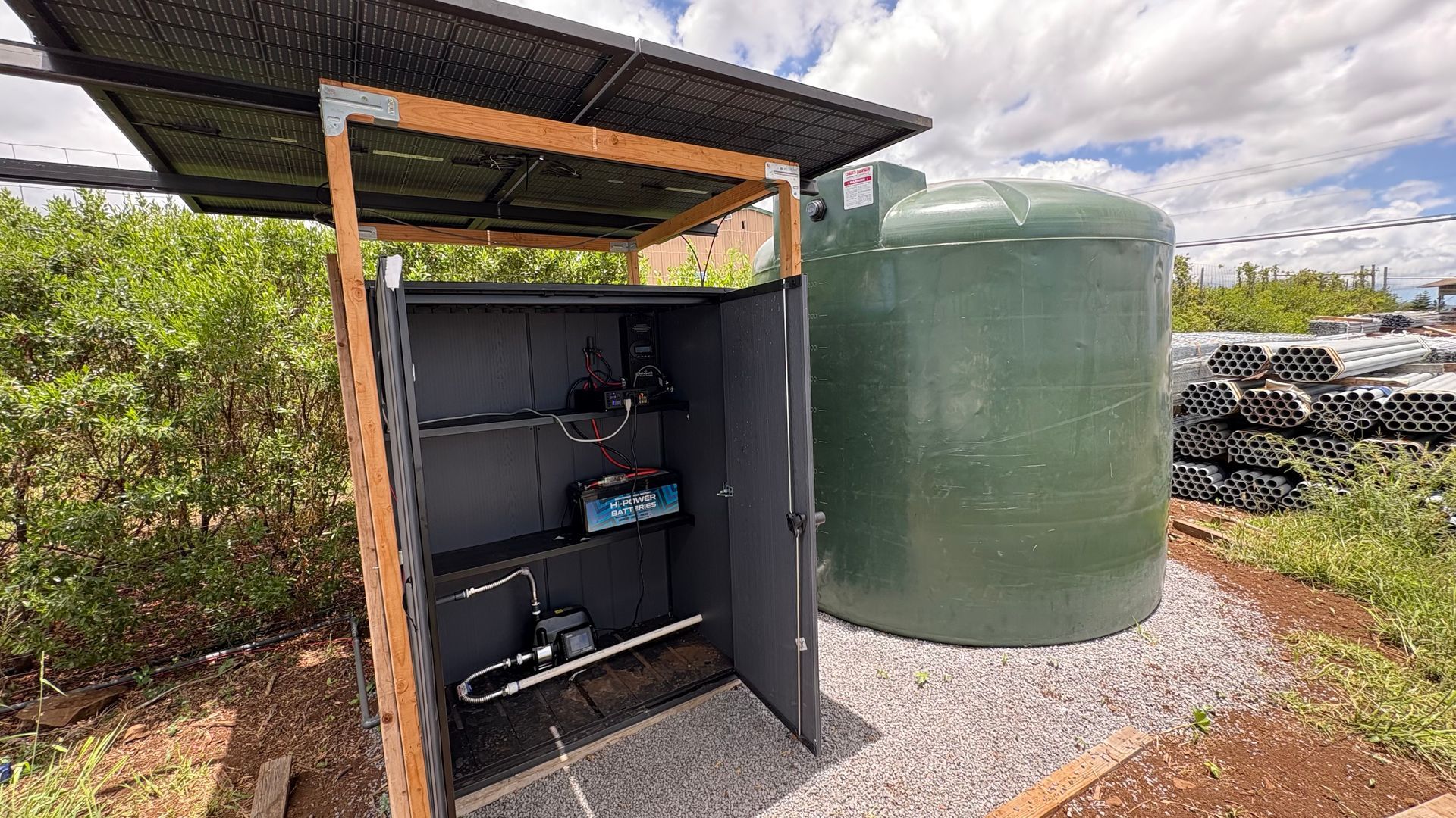 Open equipment shed with solar panel roof next to a green water tank and piles of pipes.