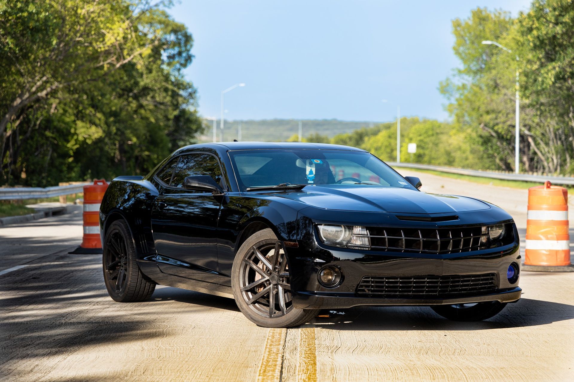 Black Chevrolet Camaro parked on road, flanked by traffic cones, trees in background.