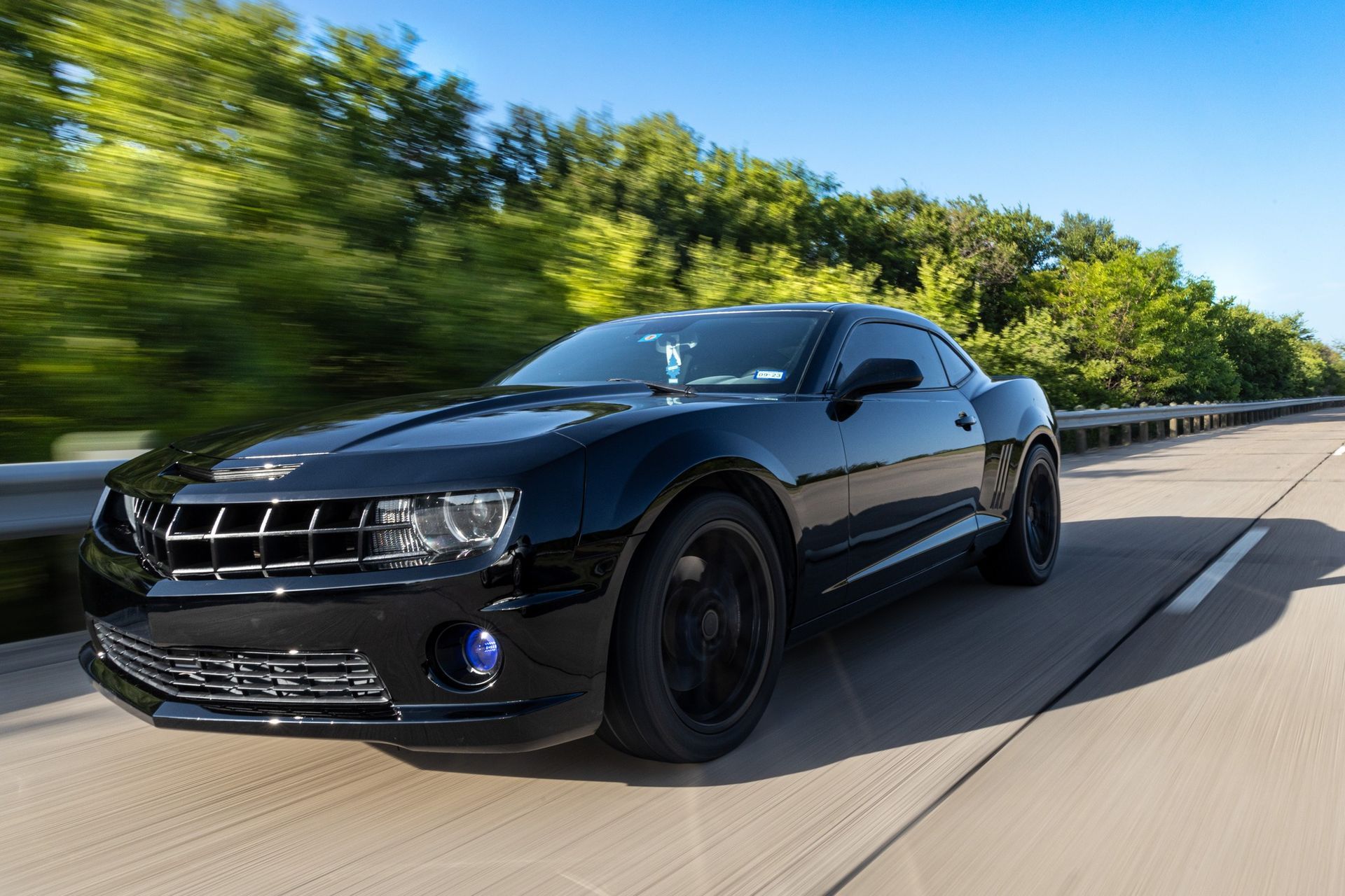 Black Chevrolet Camaro speeding on a highway with blurred green trees in the background under a blue sky.