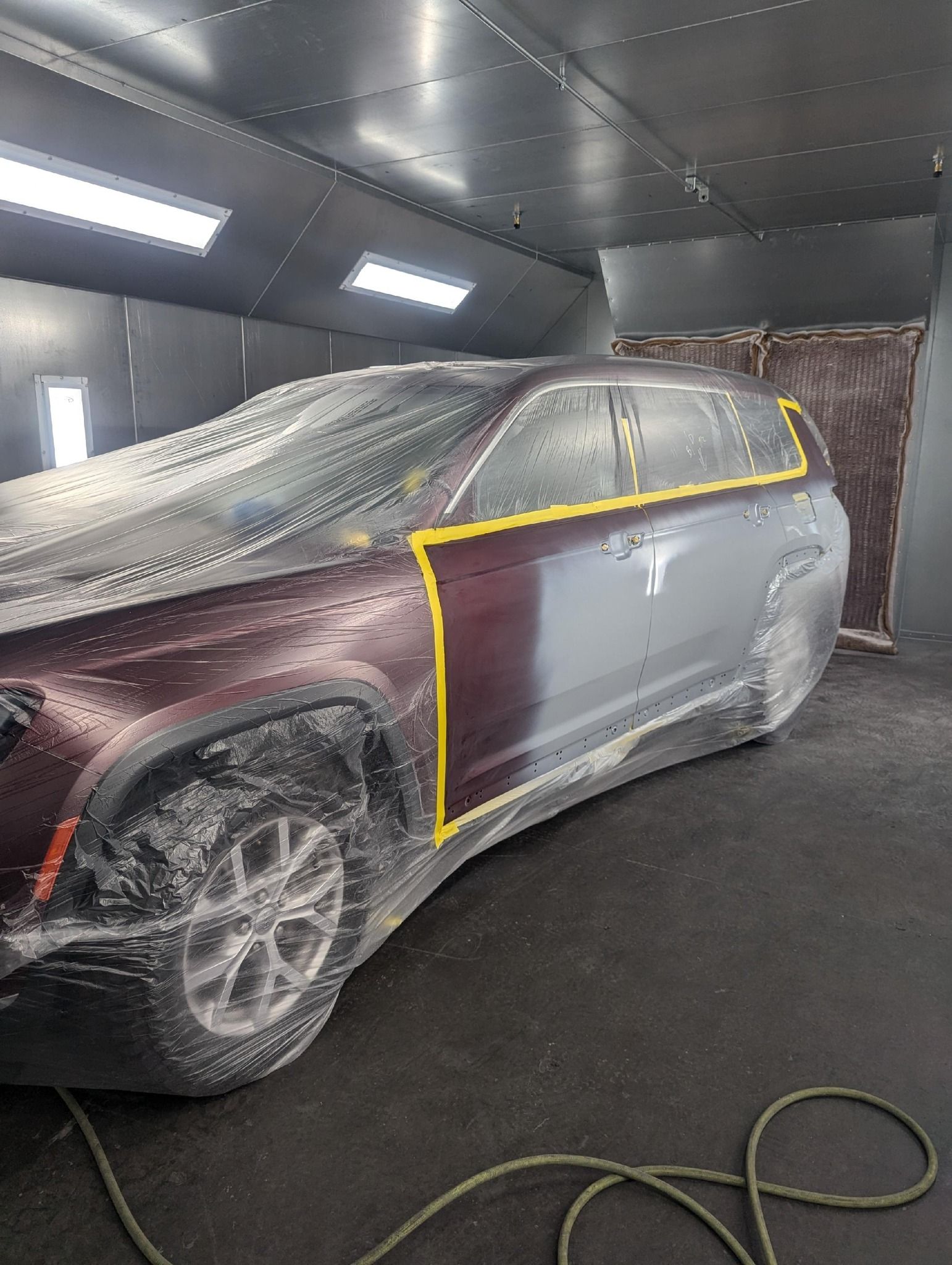 Car partially painted in a paint booth; burgundy and gray colors. Masking tape and plastic covering sections.