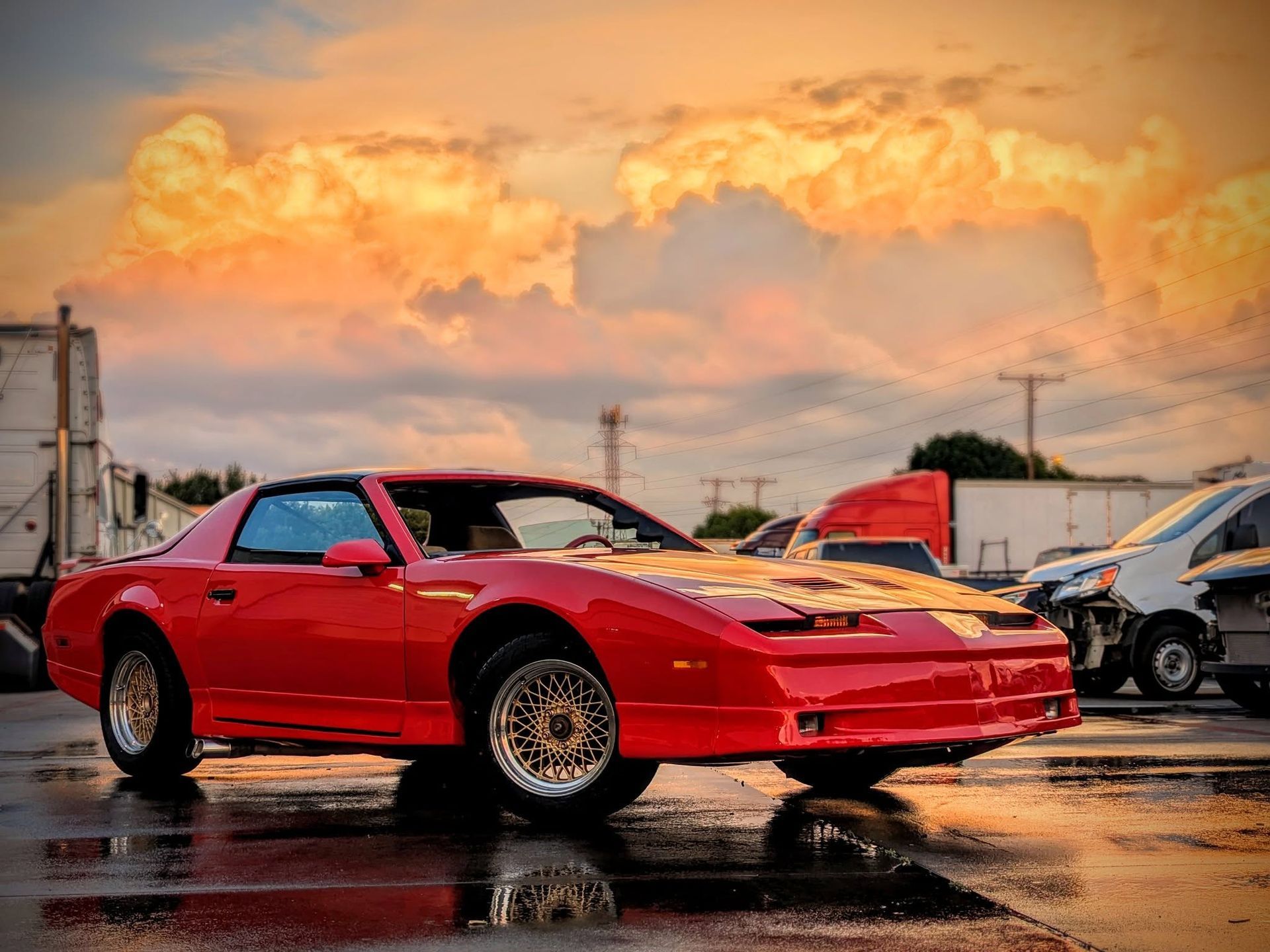 Red Pontiac Firebird car parked on wet asphalt with an orange and yellow sky in the background.