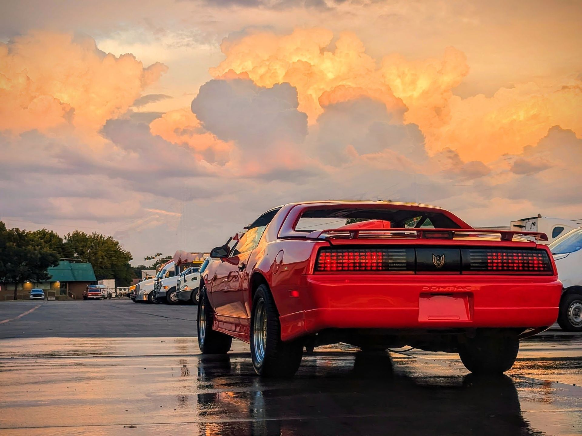 Red Pontiac Firebird on wet pavement, against a dramatic sunset sky.