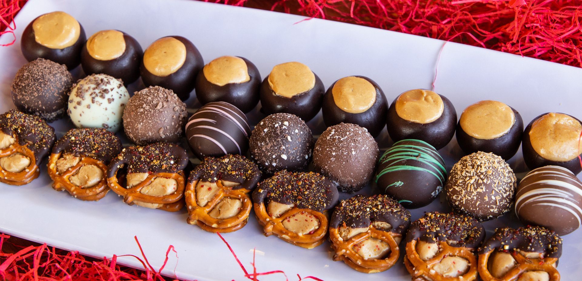 A white platter with various chocolate-covered treats, including buckeyes and pretzels, on a red surface.