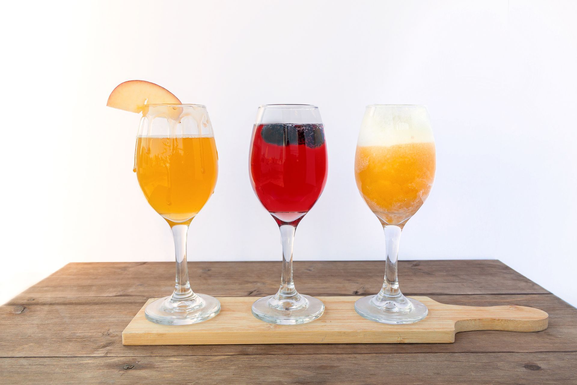 Three colorful cocktails in tall glasses on a wooden board. The board sits on a wood surface.