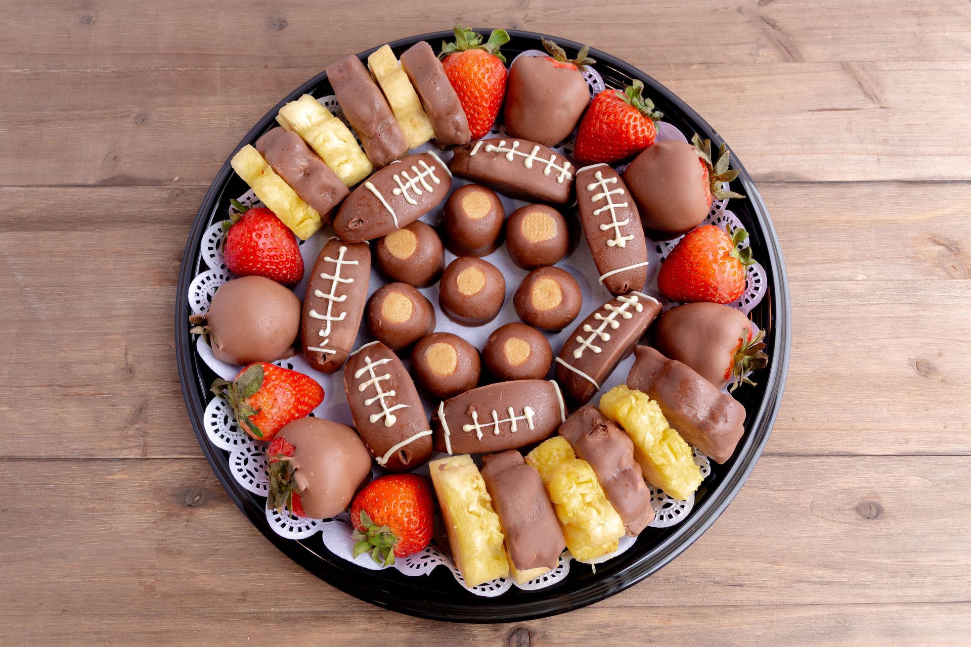 Chocolate-covered fruit platter: strawberries, pineapple, truffles, and football-shaped treats on a round, black tray.