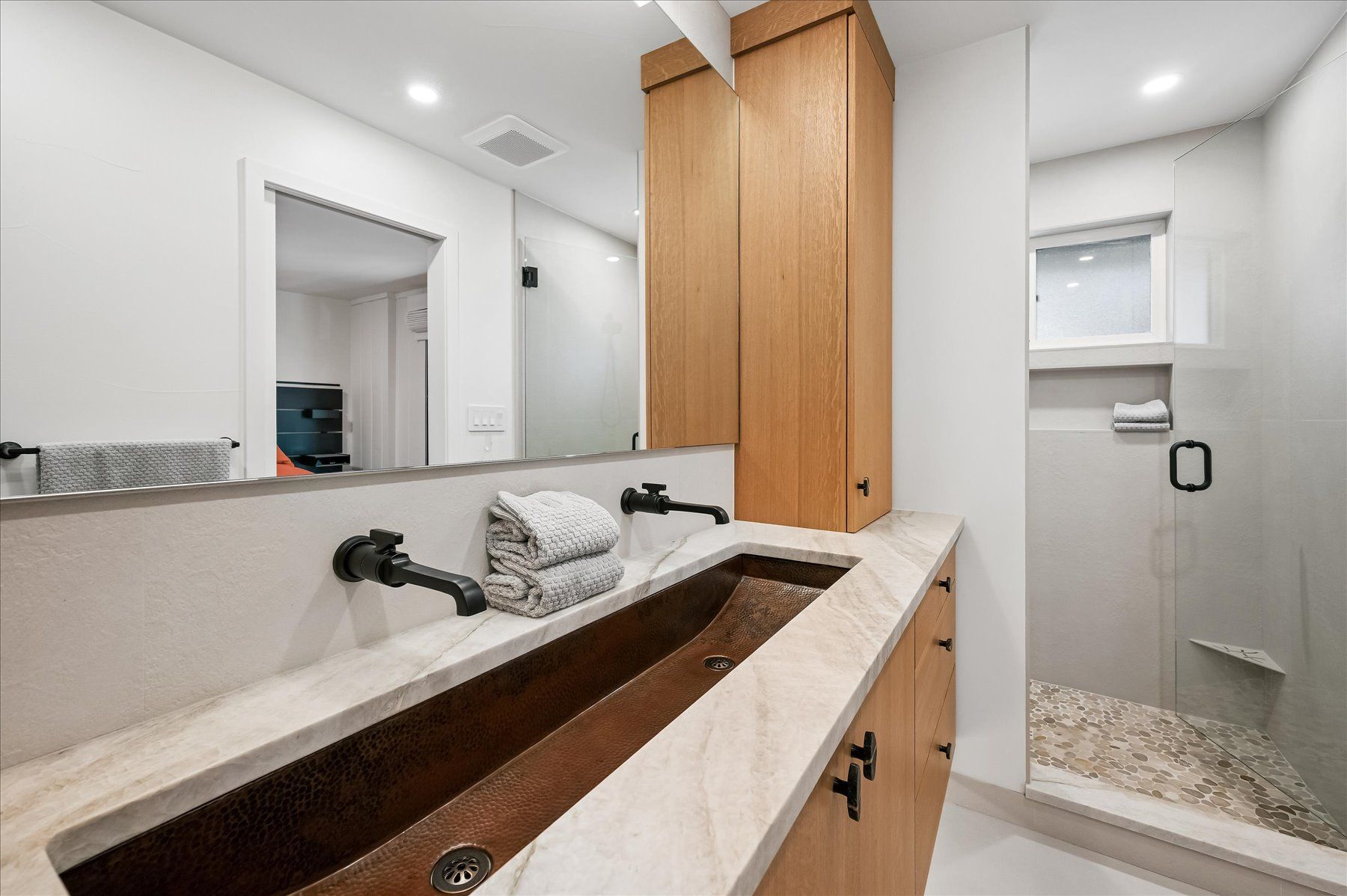 Modern bathroom vanity with long stone sink, wood cabinets, mirror, and shower stall with pebble floor.