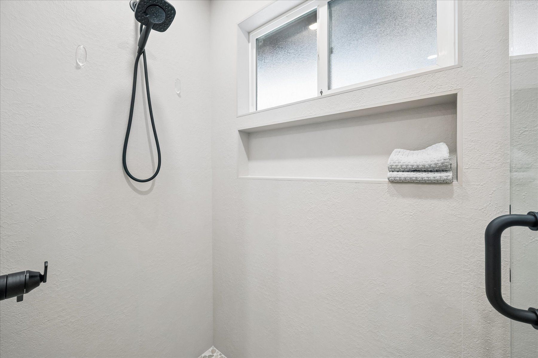 Small white shower room with black fixtures, a soap shelf, and a frosted window.