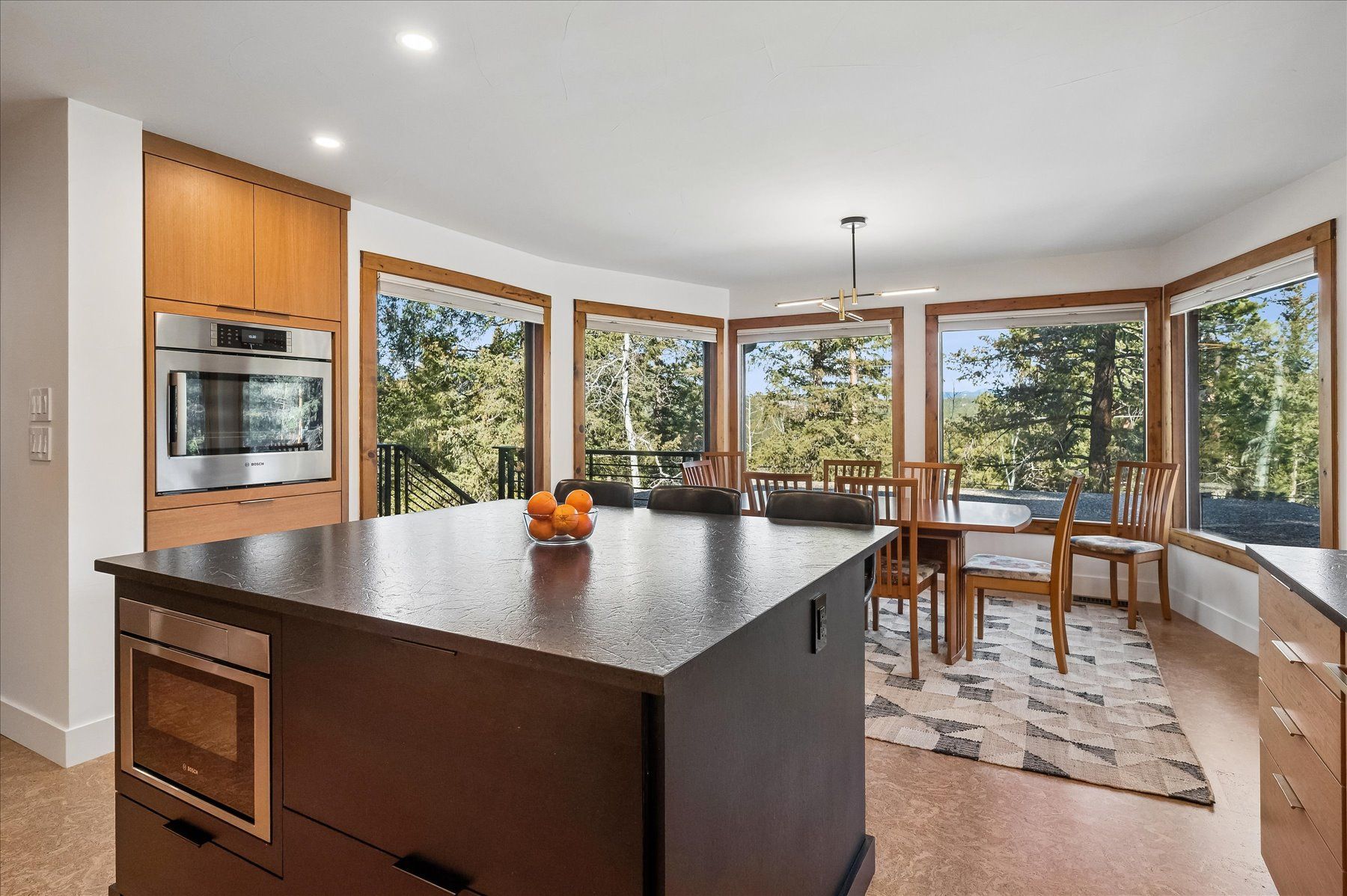 Modern kitchen with dark island, wood cabinets, and large windows overlooking trees and dining area