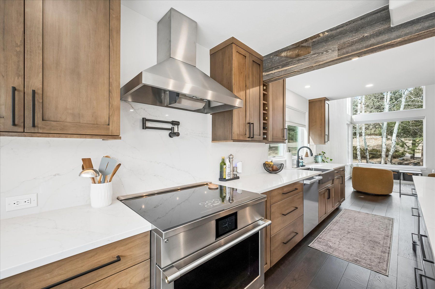 Bright modern kitchen with wood cabinets, stainless range hood, white counters, and a view into a sunlit living area