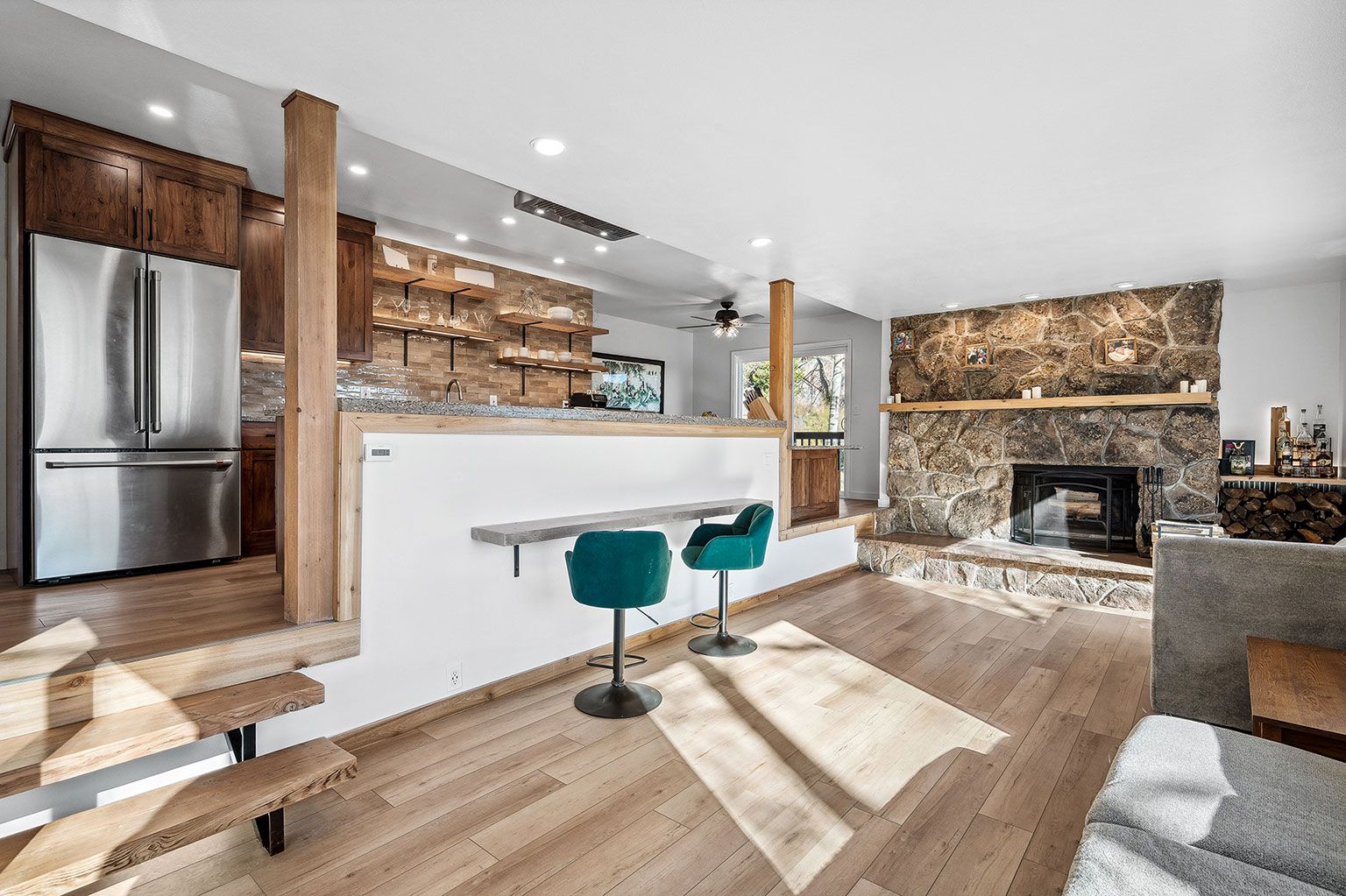 A living room with hardwood floors, a stainless steel refrigerator, a stone fireplace, and a bar.