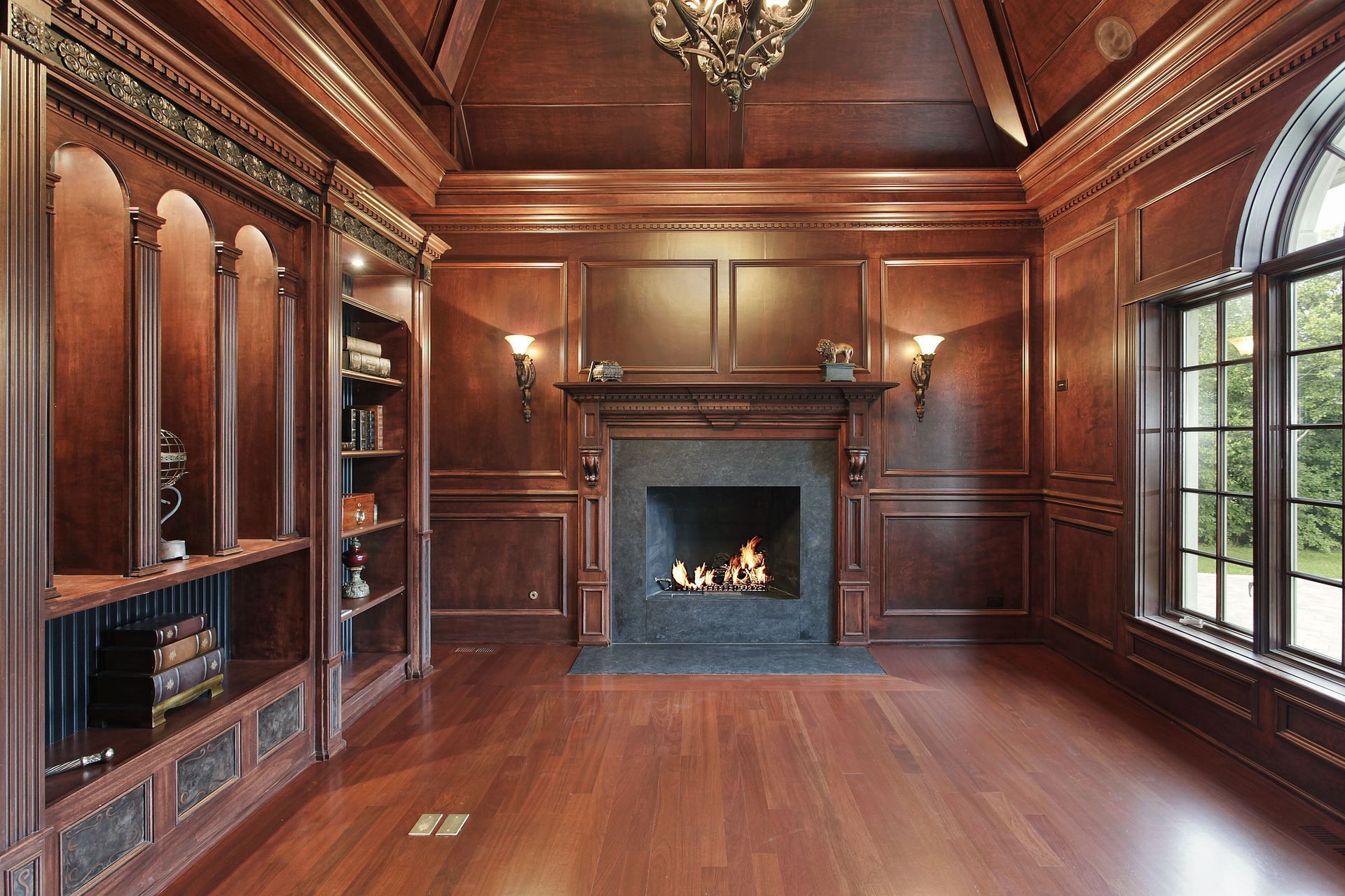 Wood-paneled library with built-in bookshelves, fireplace, and arched window, all in rich, dark tones.
