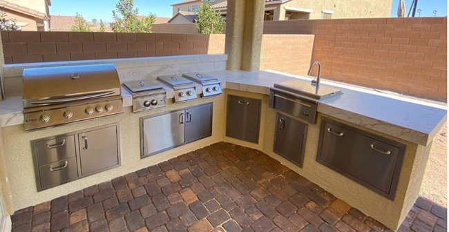 Outdoor patio with a built-in stainless steel grill and outdoor kitchen cabinets under a brick wall.