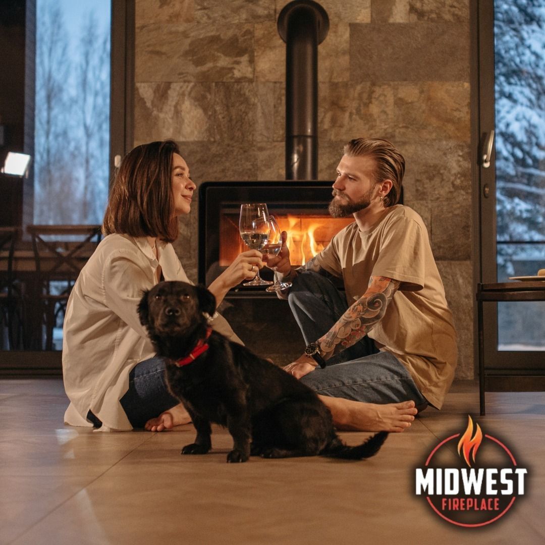 Couple toasting wine near a fireplace with their dog, seated on a wooden floor in front of a window.
