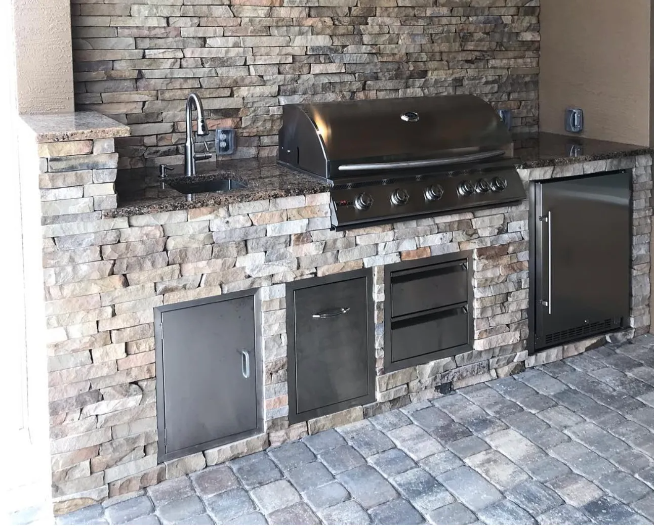 Outdoor kitchen with stone facade, stainless steel appliances, and granite countertop on a brick patio.