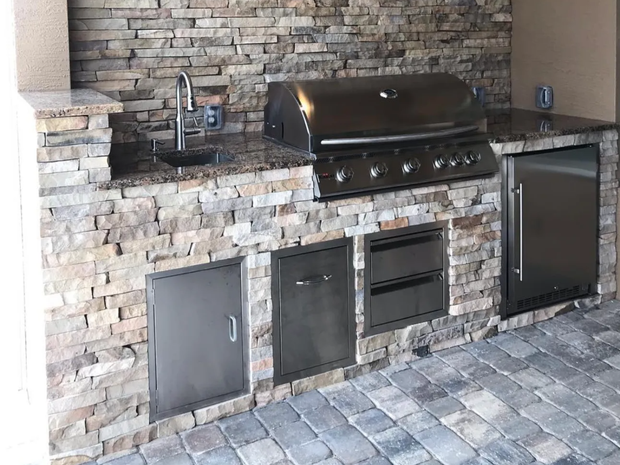 Outdoor kitchen with stone facade, stainless steel appliances, and granite countertop on a brick patio.