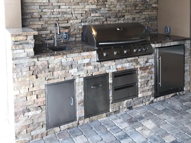 Outdoor kitchen with stone facade, stainless steel appliances, and granite countertop on a brick patio.