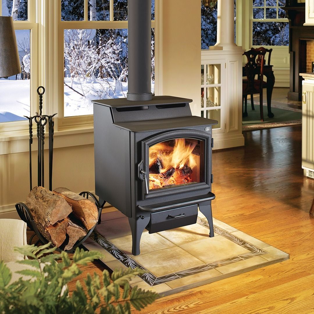 Wood-burning stove in a cozy room, lit fire, snow outside a window, wood on floor.
