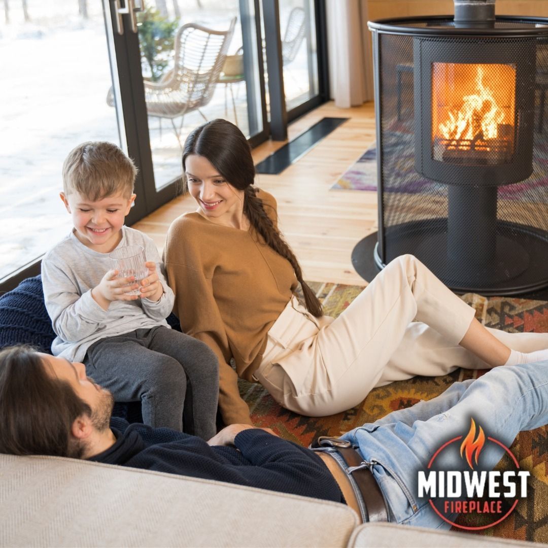 Family lounging near a fireplace with flames, wooden floor, large windows. Child holding item, smiles.
