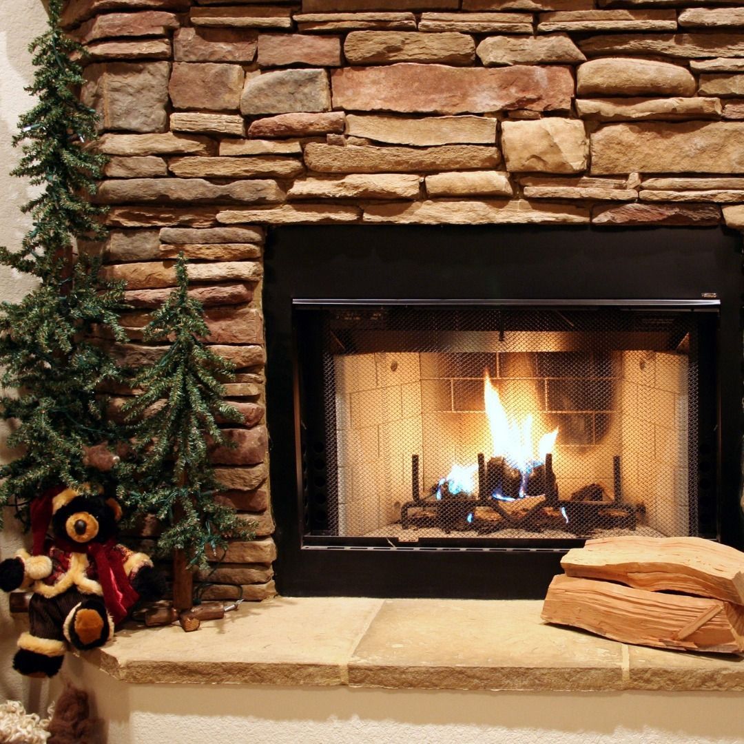 Fireplace with burning logs, stone surround, small Christmas trees, and a teddy bear.