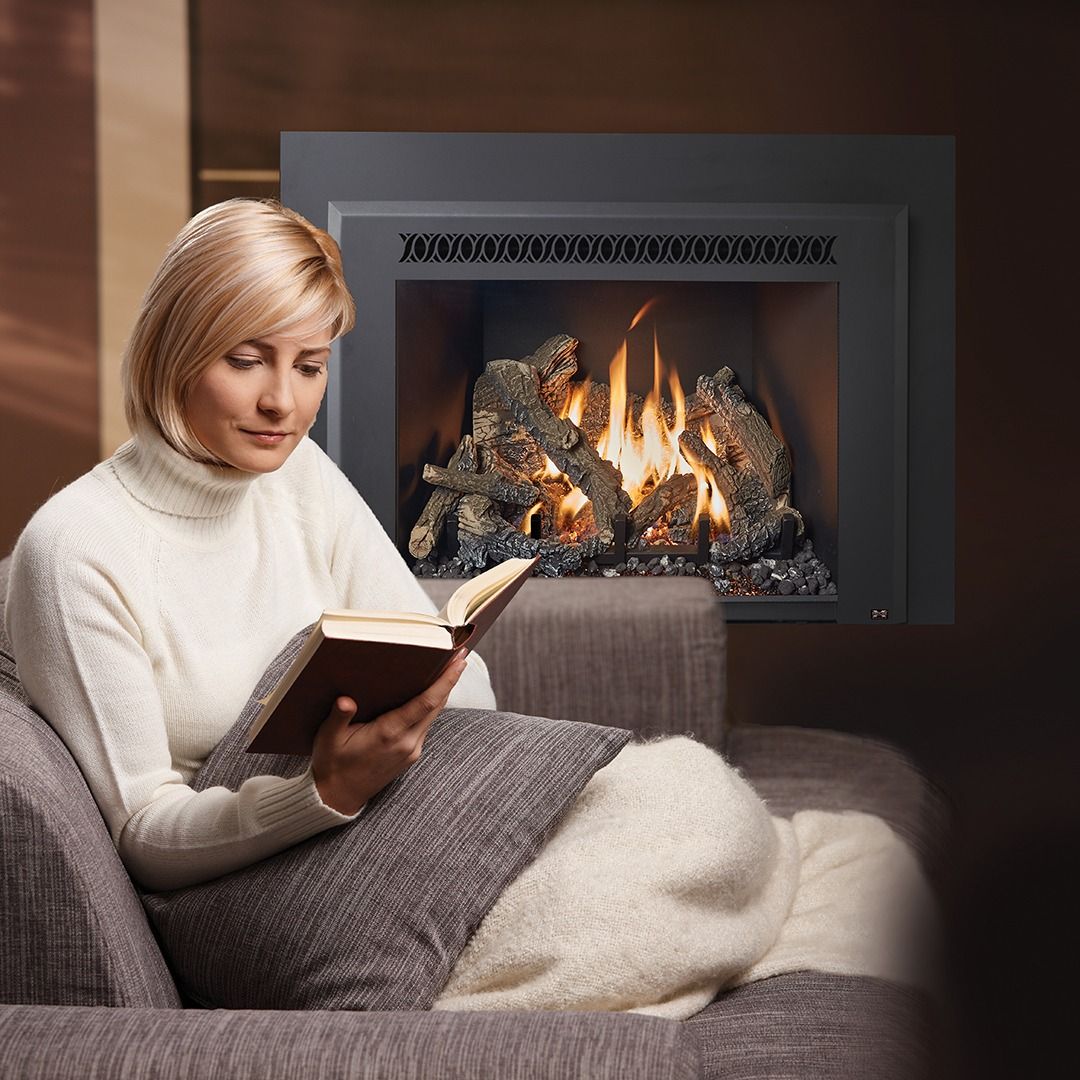 Woman reading book by a lit fireplace, sitting on a couch, wrapped in a blanket.